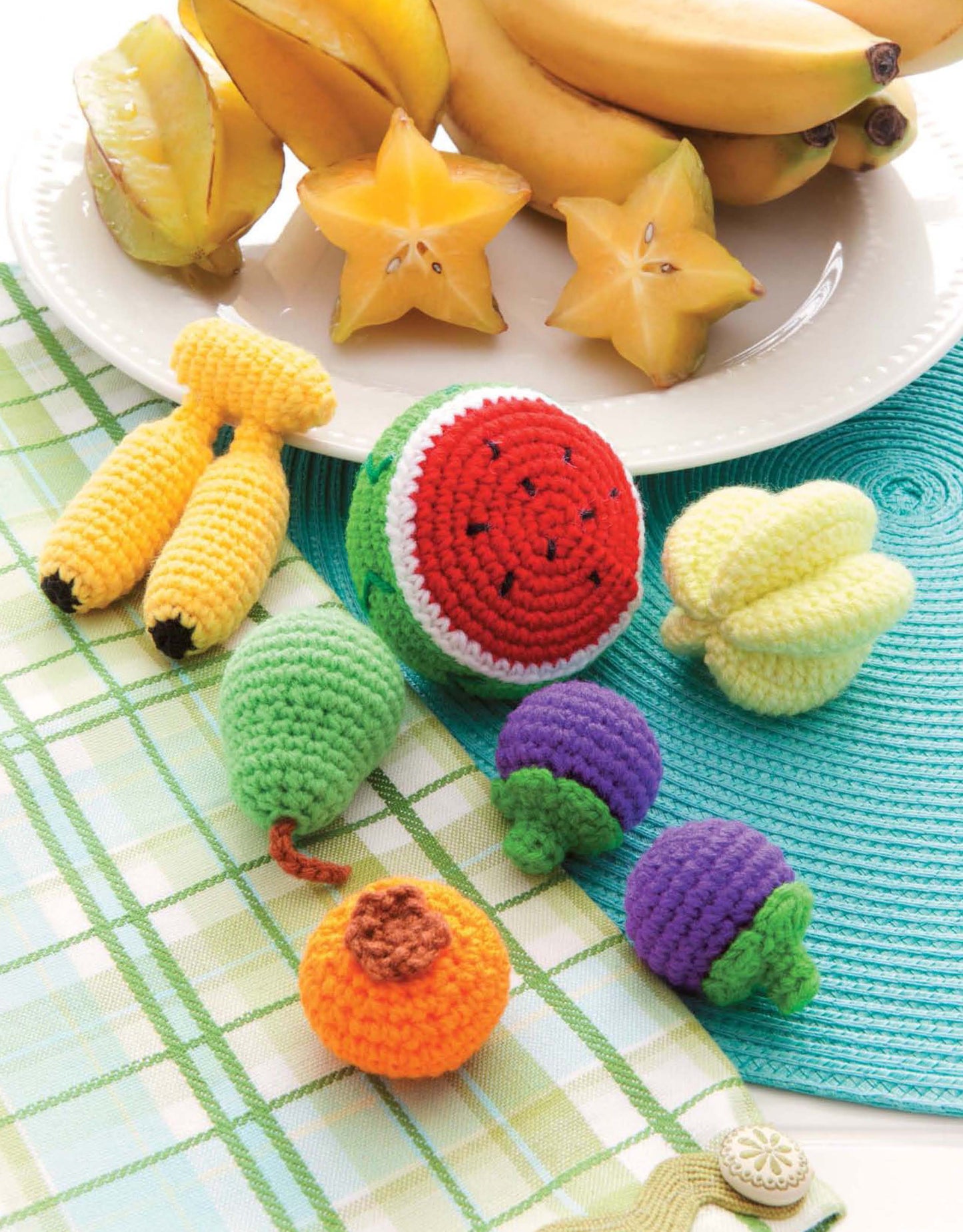 A plate with starfruit and bananas sits behind assorted crocheted fruits and vegetables—amigurumi toys like bananas, watermelon, and more—all made using the "Ice Box Crochet" pattern from Leisure Arts, arranged on a plaid and turquoise cloth.