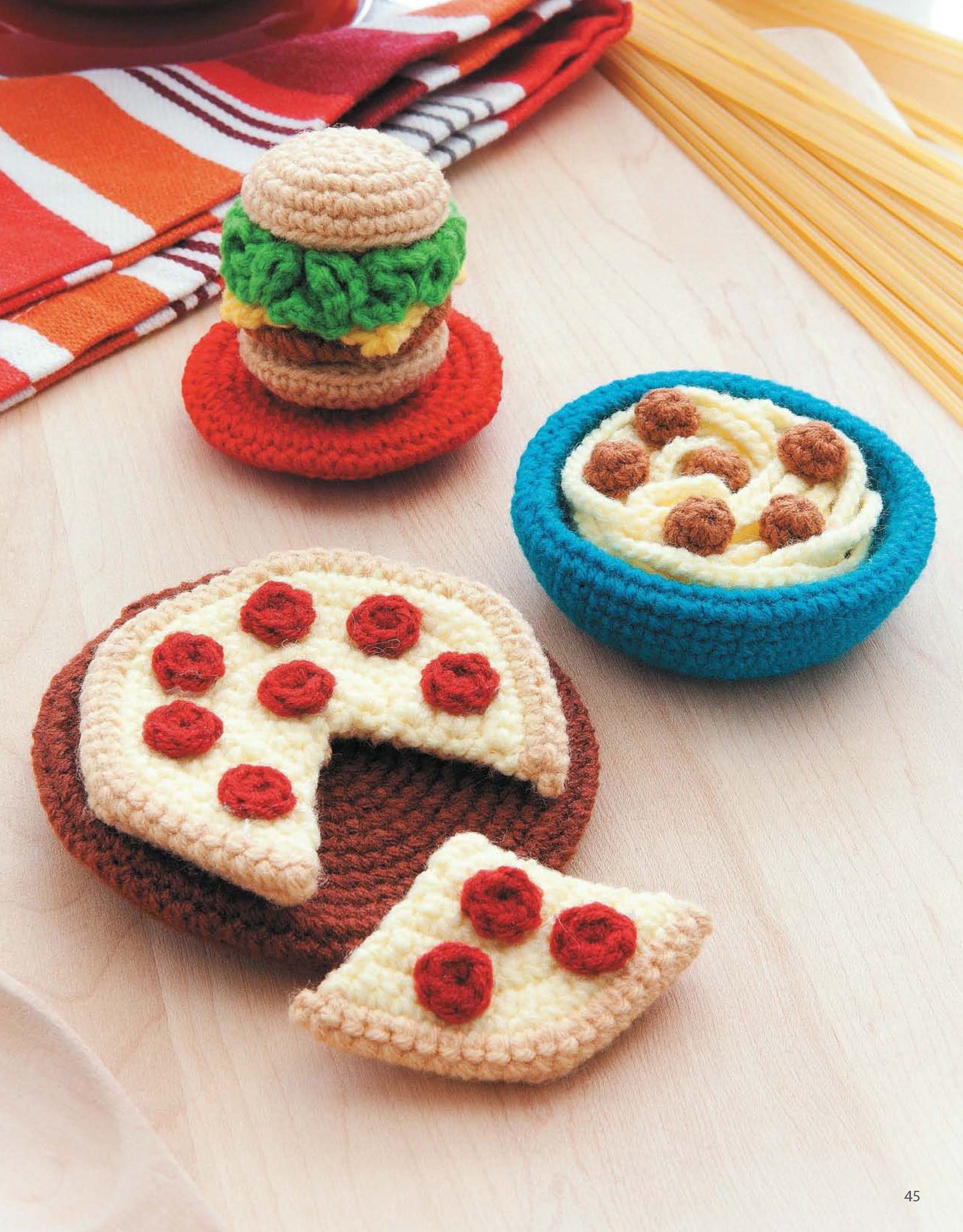 Ice Box Crochet by Leisure Arts displays charming amigurumi foods—a crocheted hamburger, a bowl of spaghetti and meatballs, and a pepperoni pizza with one slice missing—arranged on light wood near uncooked spaghetti.