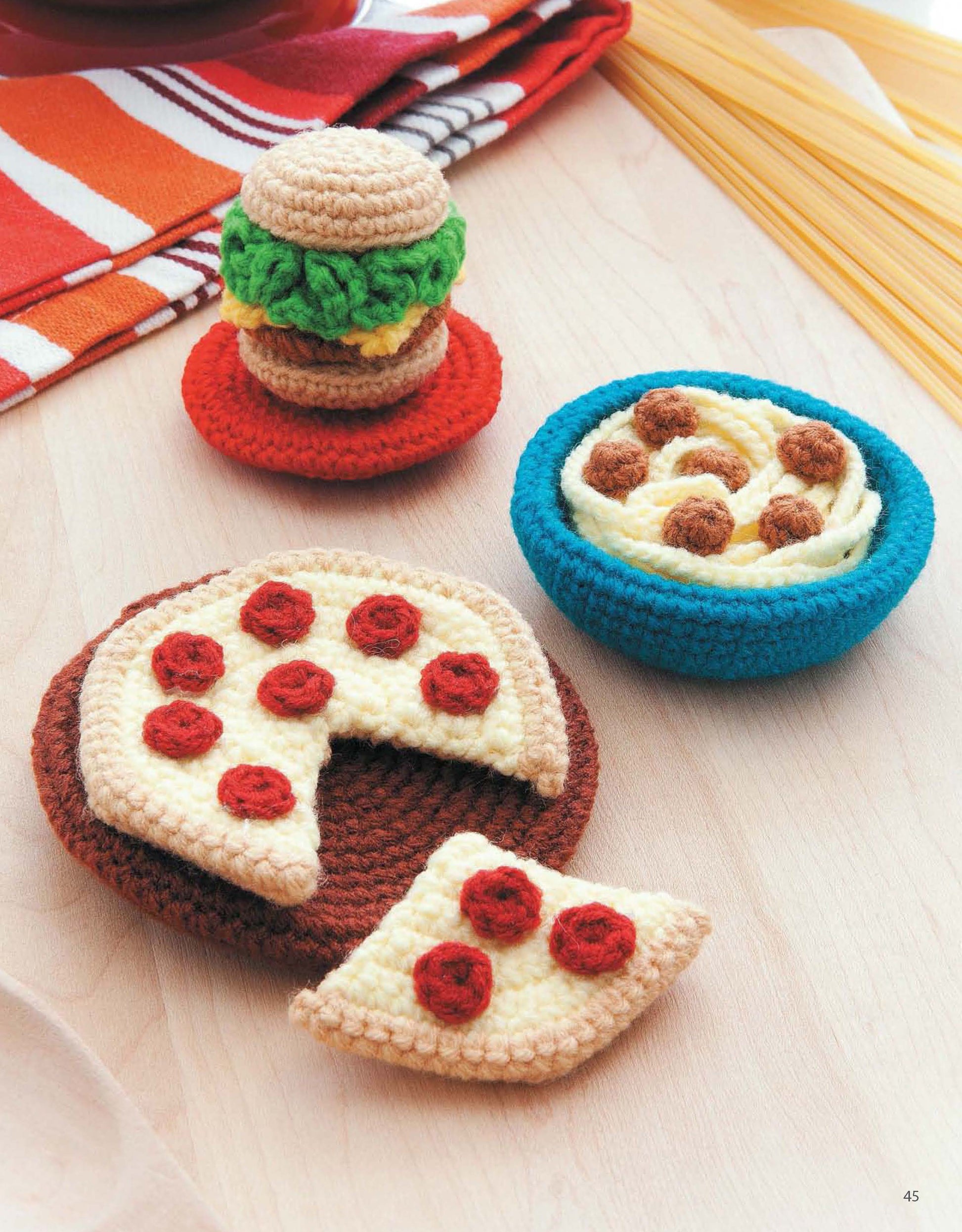 Ice Box Crochet by Leisure Arts displays charming amigurumi foods—a crocheted hamburger, a bowl of spaghetti and meatballs, and a pepperoni pizza with one slice missing—arranged on light wood near uncooked spaghetti.