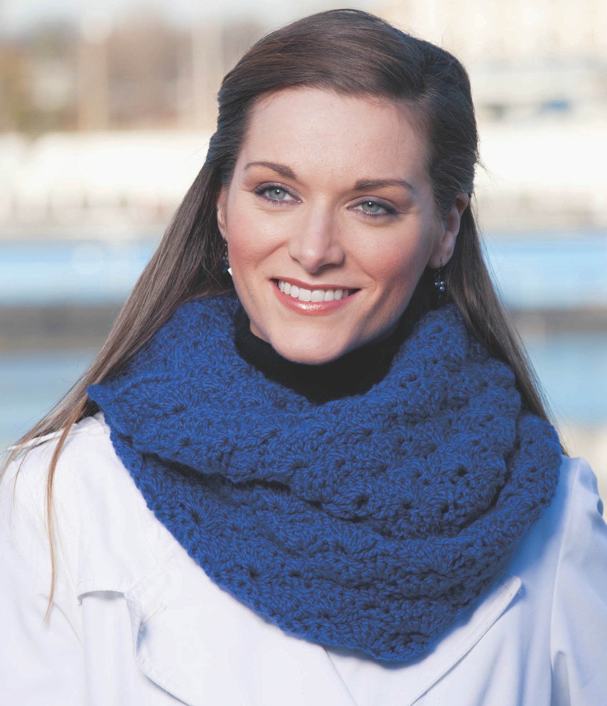 A woman with long brown hair smiles outdoors, wearing a white coat and a cobalt blue Crochet Cowl by Leisure Arts, inspired by easy crochet designs, with water and buildings blurred in the background.