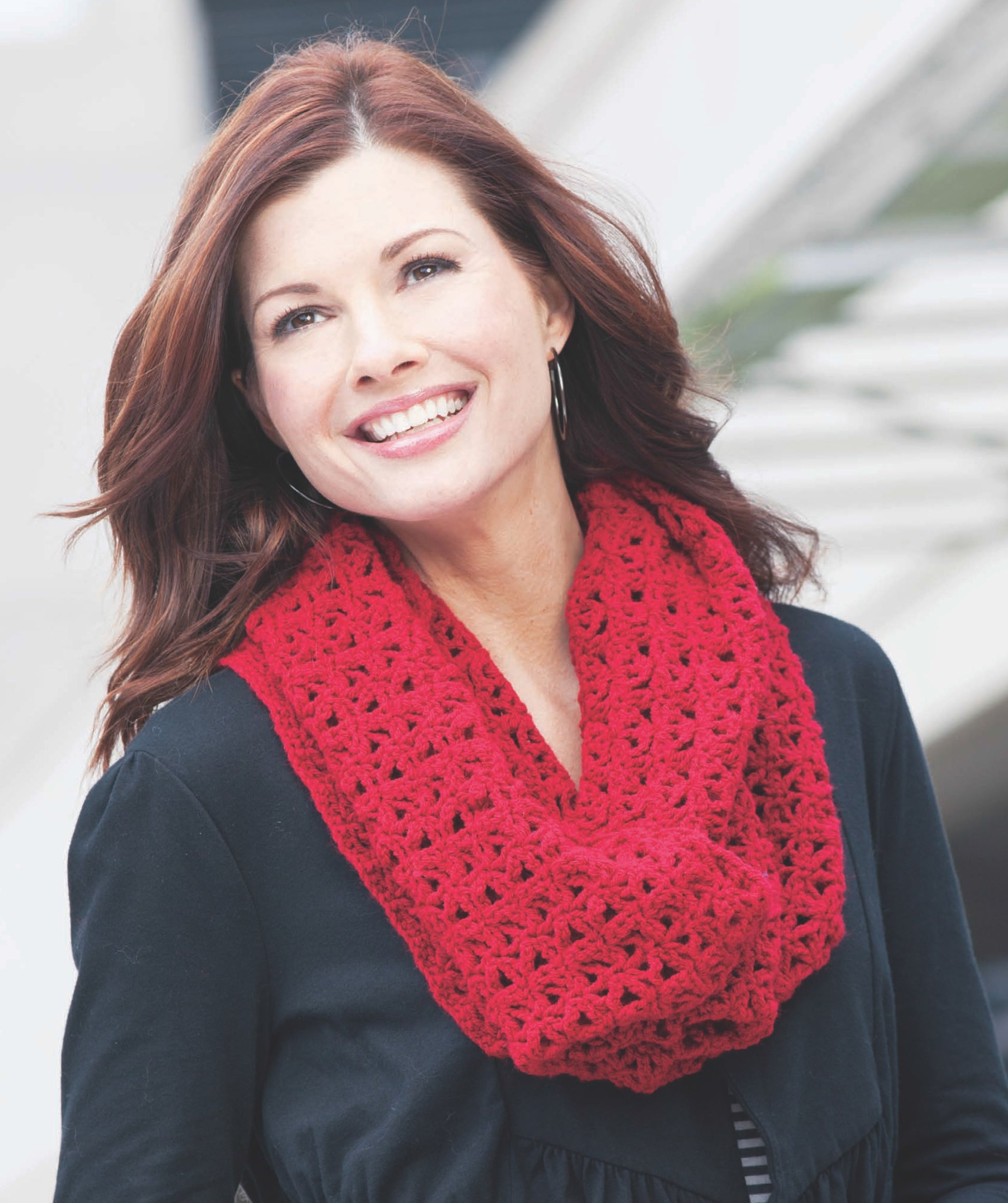 A woman with shoulder-length brown hair smiles outdoors in natural light, wearing a bright red Crochet Cowl from Leisure Arts over a black coat.