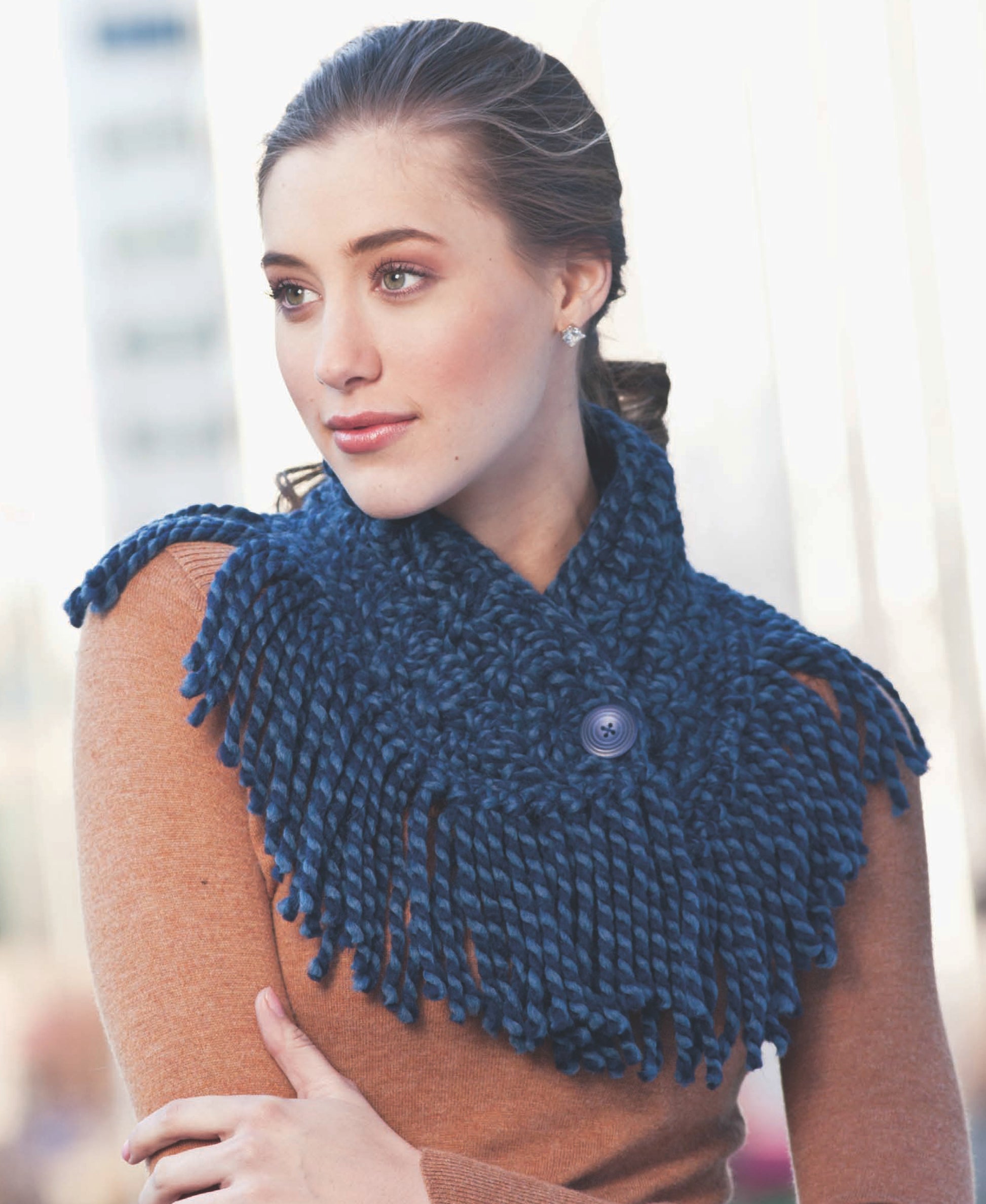 A woman models a chunky, dark blue Crochet Cowl by Leisure Arts with fringe and a large button—an inspiring accessory for easy crochet designs, set against a softly blurred urban background.