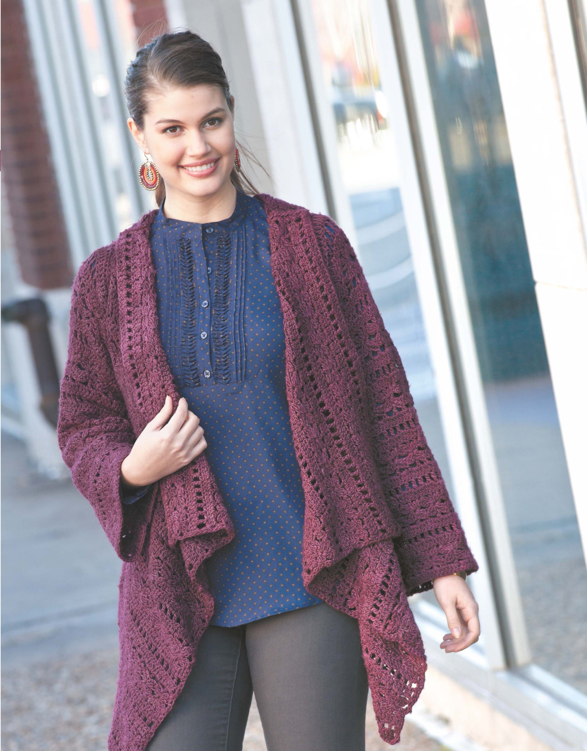 A woman smiles outside in a loose, dark purple crochet cardigan from Cool Weather Fashions by Leisure Arts, layered over a blue shirt and dark pants. Her brown hair is tied back, and she completes the look with bold red earrings.