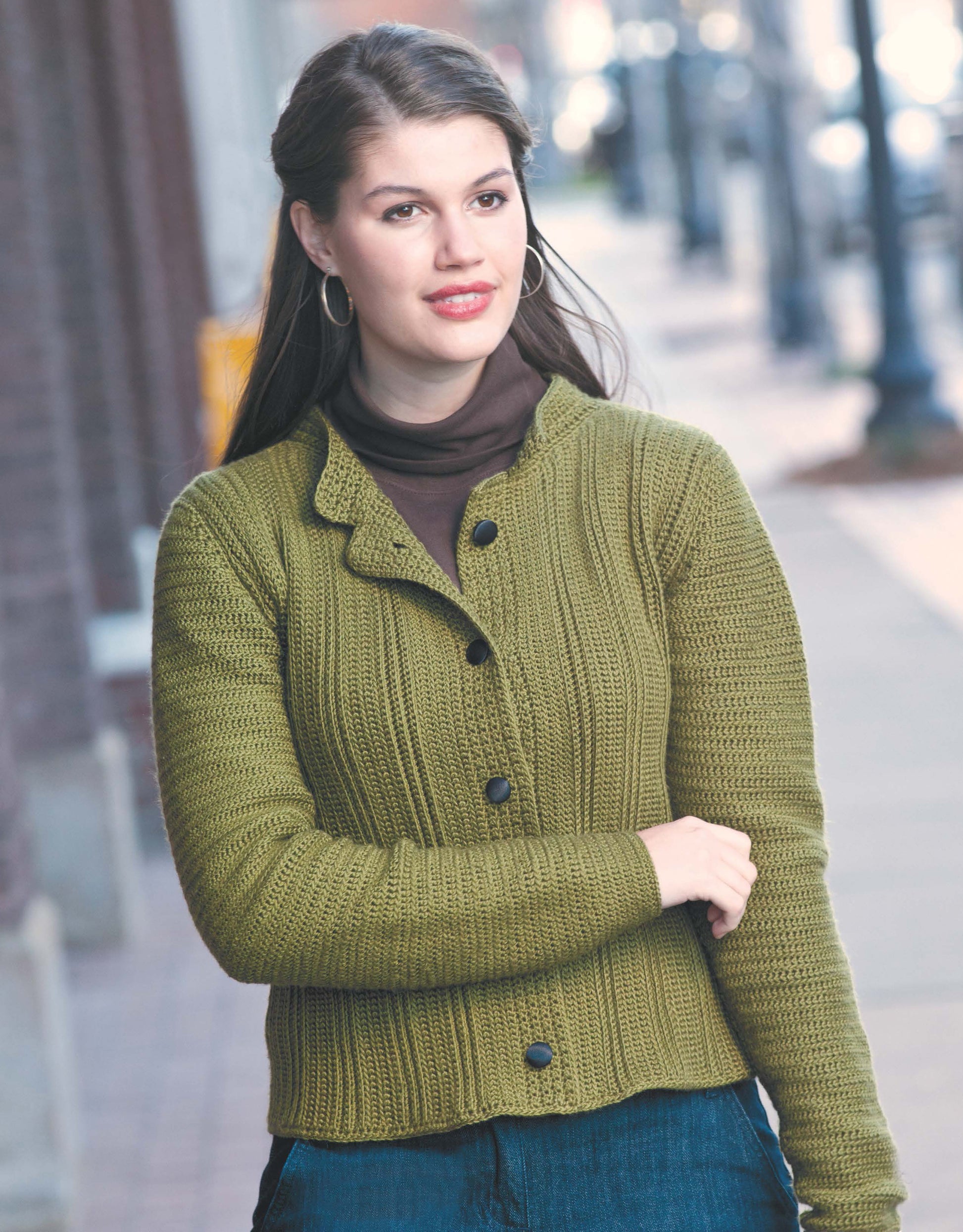 A woman wearing Cool Weather Fashions by Leisure Arts—a brown turtleneck and olive green ribbed crochet cardigan—stands on a city sidewalk with blurred buildings and streetlights in the background.