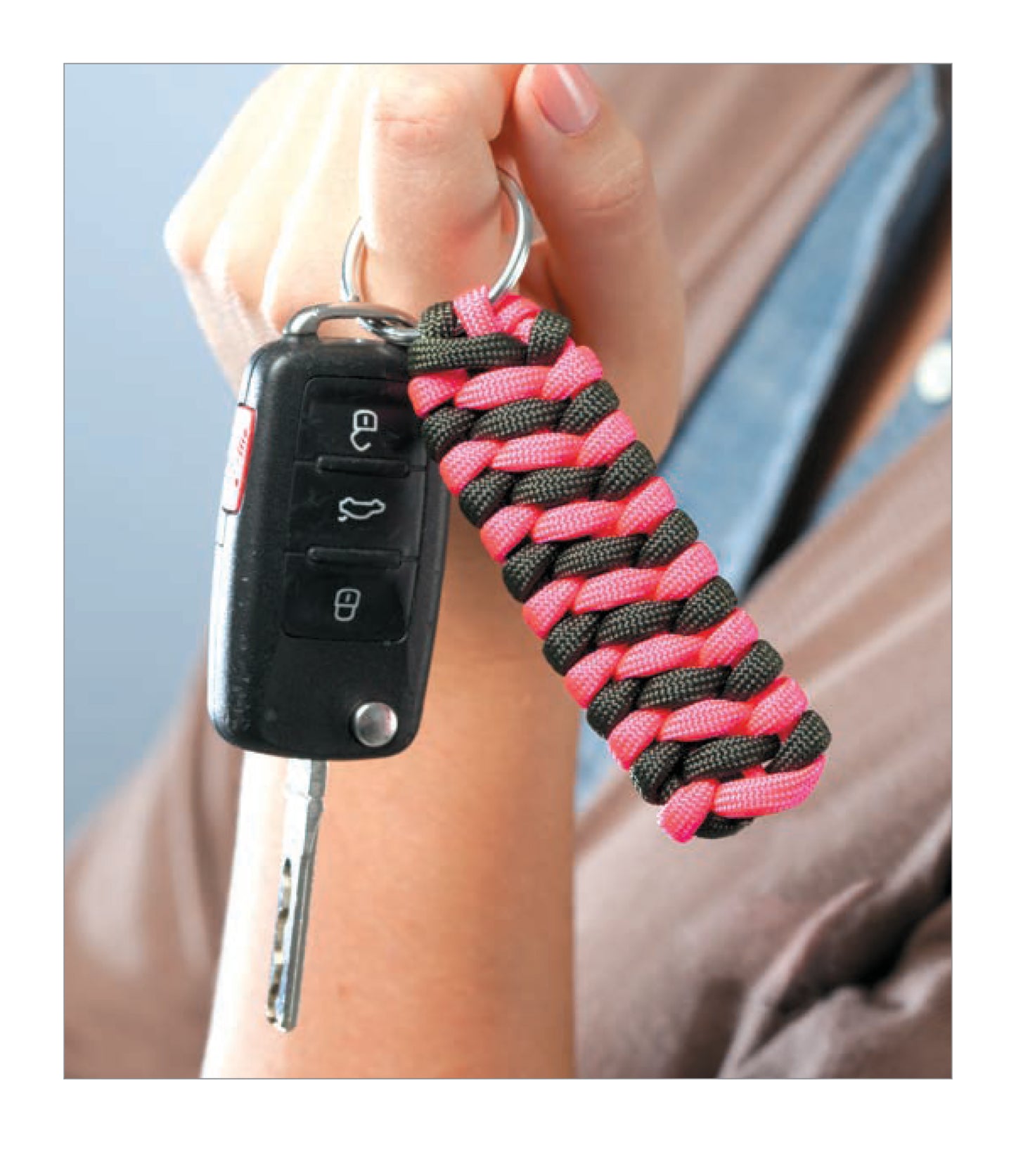 A hand holds a car key with a black remote and a pink-and-black DIY Jewelry Paracord keychain from Leisure Arts, ideal for beginners practicing simple bracelet knot techniques.