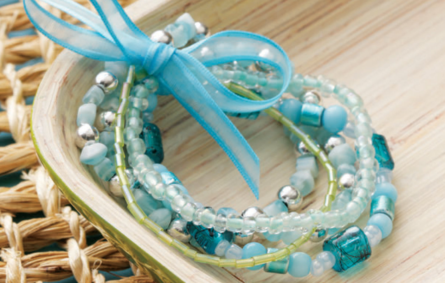 DIY Jewelry Learn the Basics by Leisure Arts features blue and silver beaded bracelets tied with a sheer blue ribbon, displayed on a wooden tray—ideal for beginner jewelry makers looking for easy projects.
