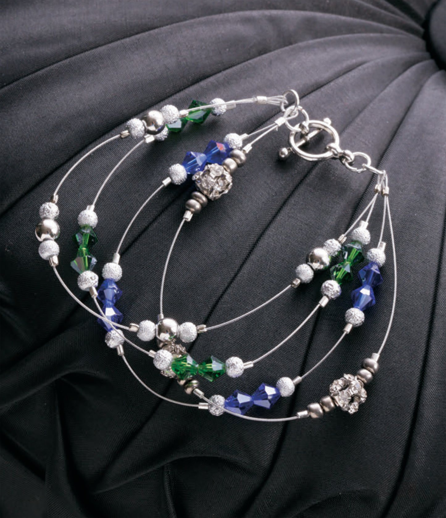 Leisure Arts DIY Jewelry Learn the Basics features a multi-strand silver bracelet with blue, green, and silver crystal and metallic beads—shown on black pleated fabric—ideal for beginner jewelry makers seeking easy projects.