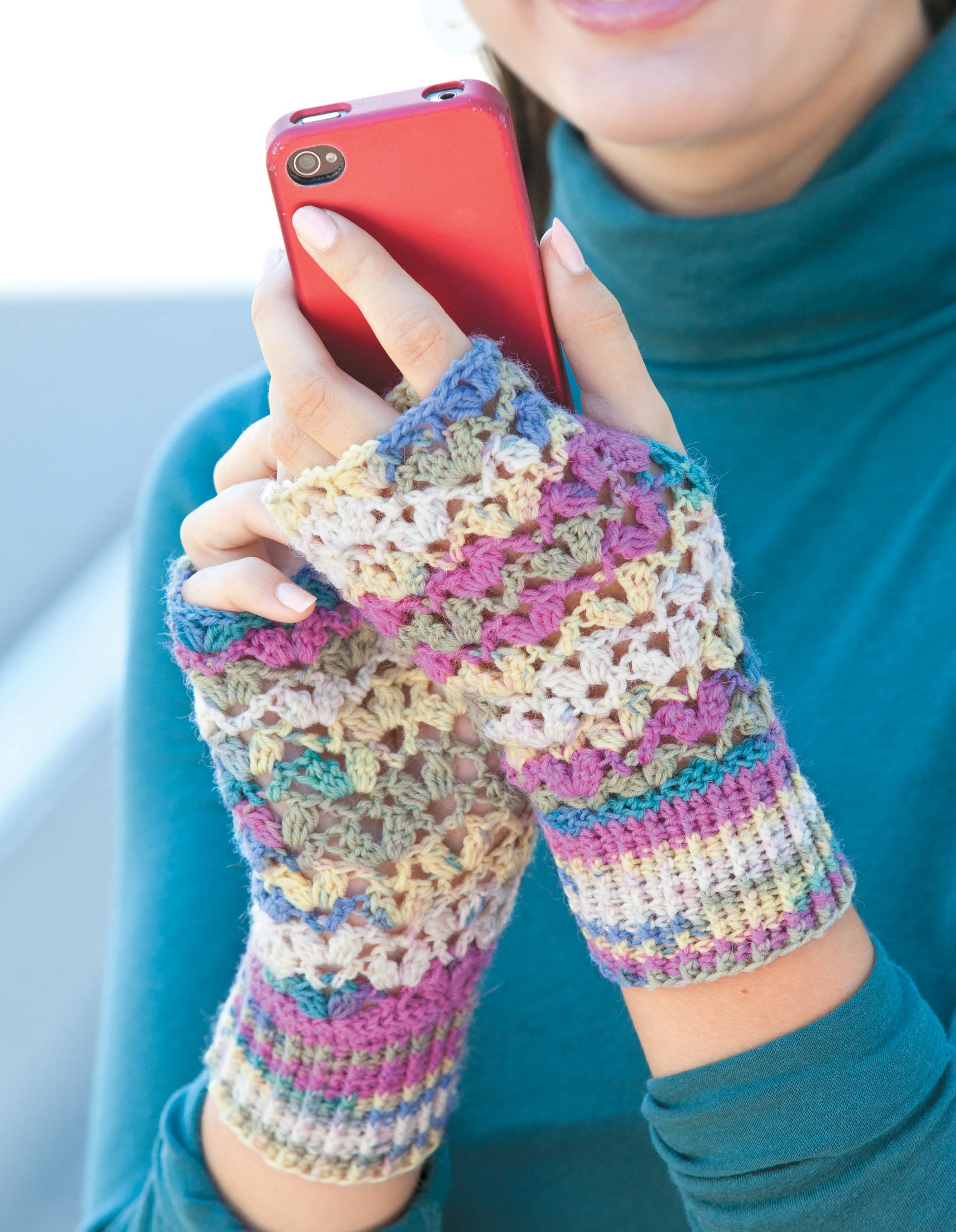 A person in a teal turtleneck smiles while holding a red phone, showing off colorful Texting Mitts—Stay Toasty Warm With Sock-Yarn Mitts! by Leisure Arts. The vibrant crocheted fingerless gloves are perfect for keeping hands warm while using devices.