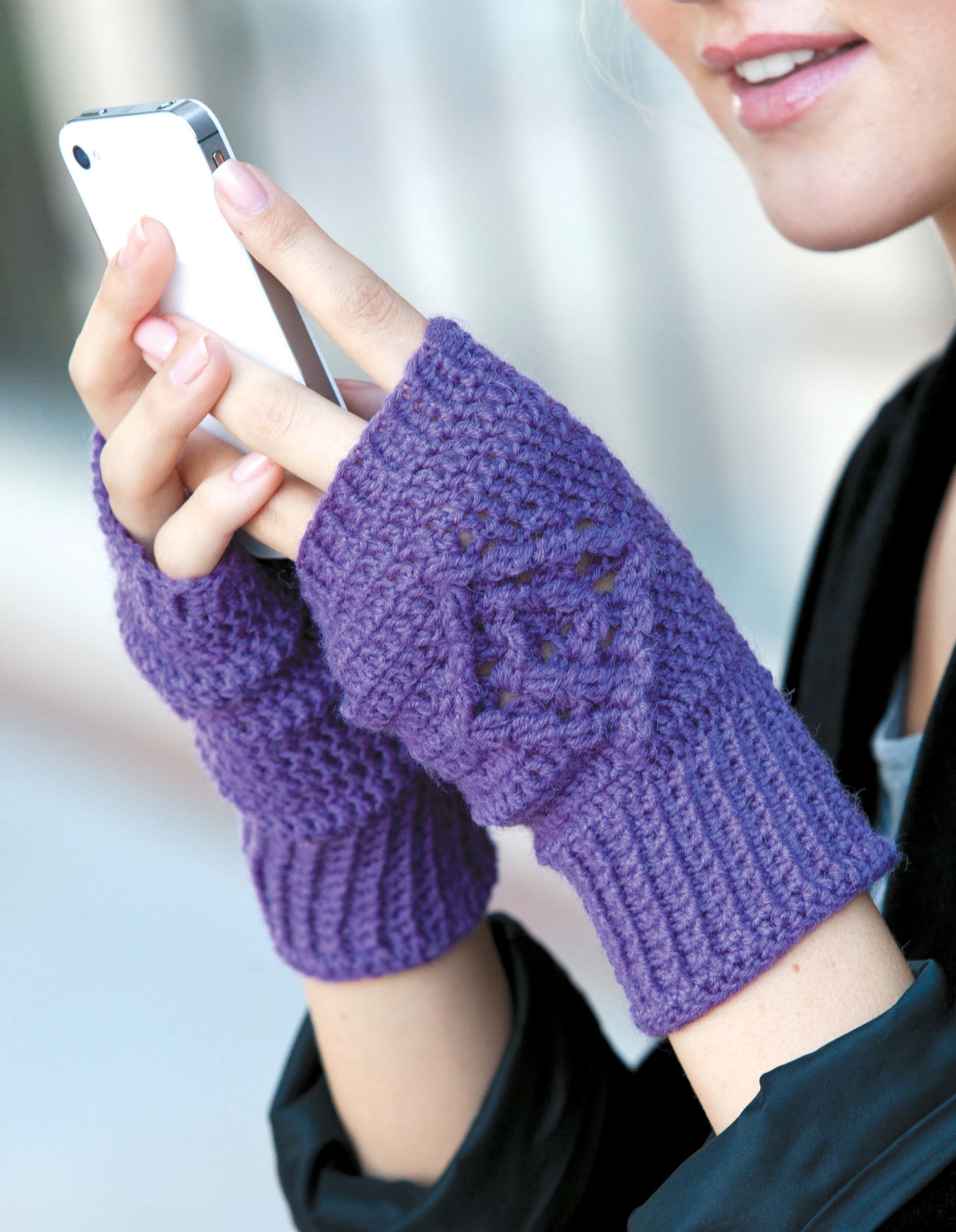 Someone wearing Leisure Arts' Texting Mitts—Stay Toasty Warm With Sock-Yarn Mitts! is using a smartphone, smiling softly. Only the lower face and hands are shown.