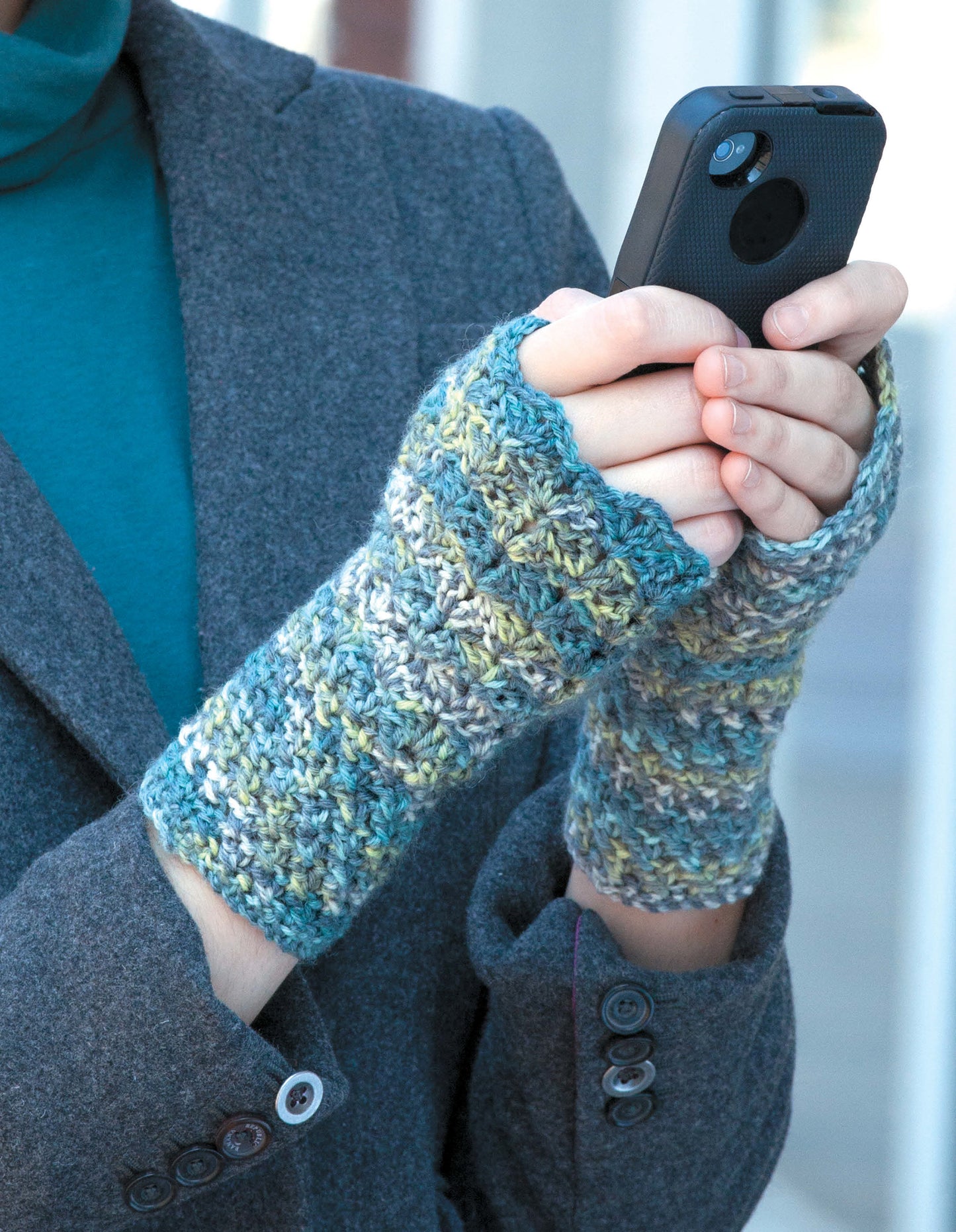 A person wearing Leisure Arts' Texting Mitts—Stay Toasty Warm With Sock-Yarn Mitts! in blue-green and a gray coat holds a smartphone with both hands outdoors, the soft, hand-knit fingerless gloves perfect for texting.