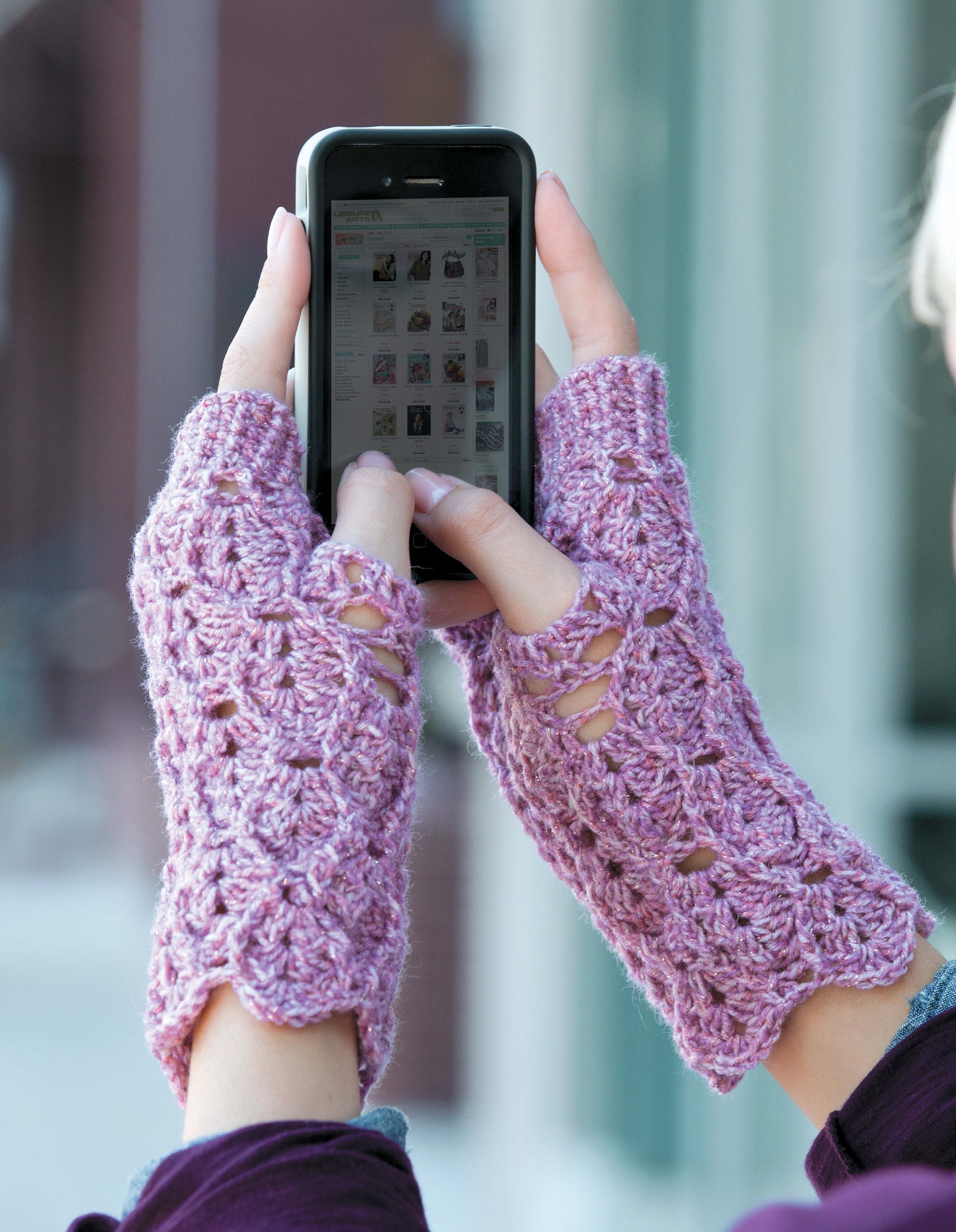 A person outdoors near a glass building uses a smartphone while wearing pink Texting Mitts—Stay Toasty Warm With Sock-Yarn Mitts! by Leisure Arts, showcasing their intricate crocheted fingerless design.