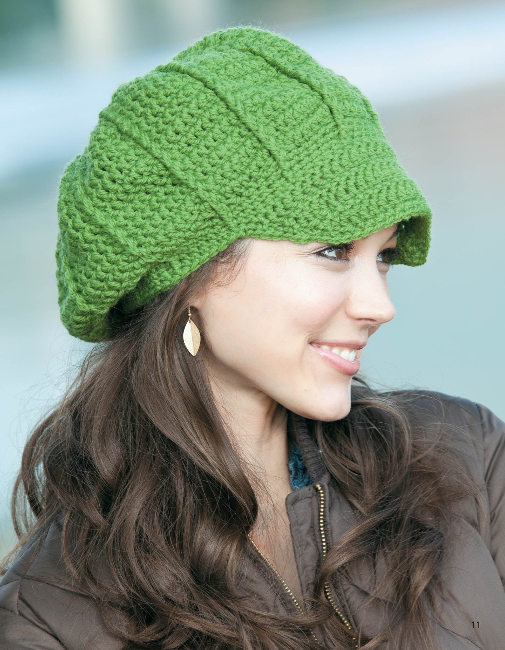 A woman with long brown hair wears a bright green hat made from the "Slouchy Beanies and Headwraps Digital Download" by Leisure Arts, paired with gold leaf-shaped earrings and a brown jacket, smiling against a blurred outdoor background.