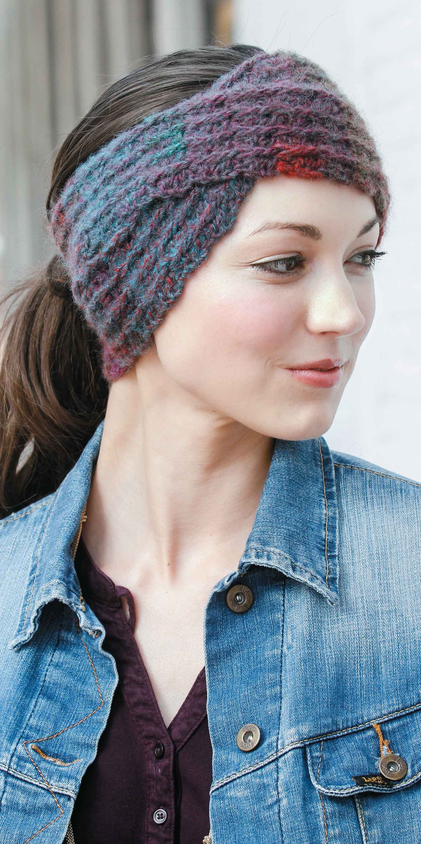 A woman with dark hair pulled back models a colorful headwrap from the Leisure Arts Slouchy Beanies and Headwraps Digital Download, paired with a denim jacket and dark shirt, giving inspiration for beginner crocheters.