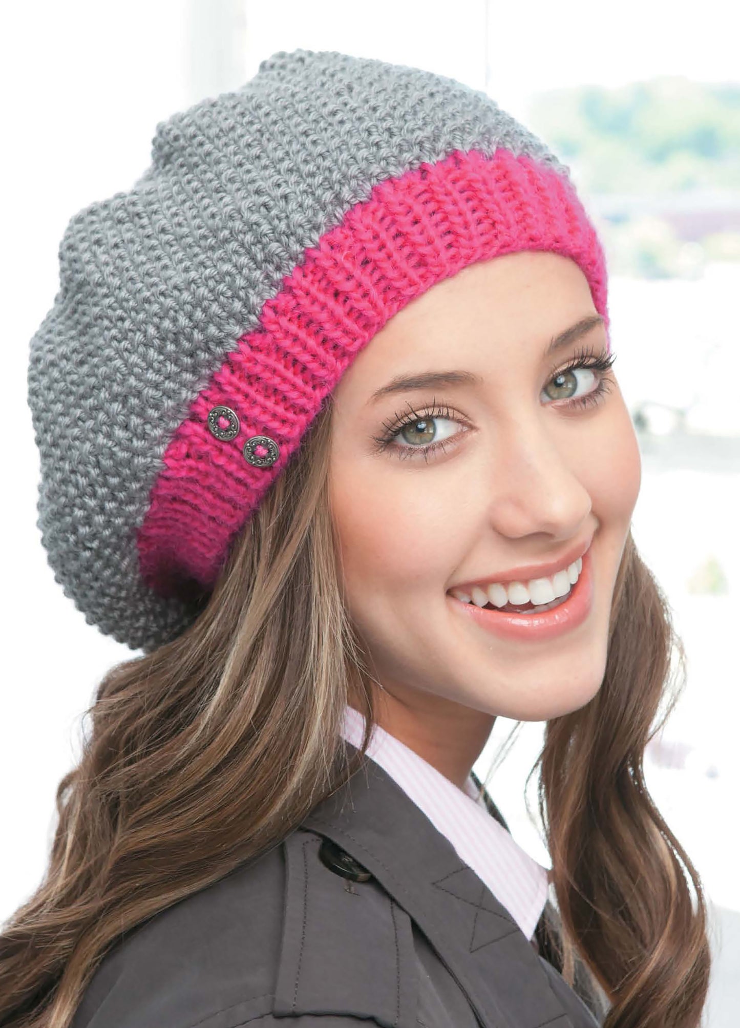 A young woman with long brown hair smiles while wearing a stylish gray knit beret with a bright pink band from Leisure Arts' Slouchy Beanies and Headwraps Digital Download, paired with a gray jacket against a softly blurred background.