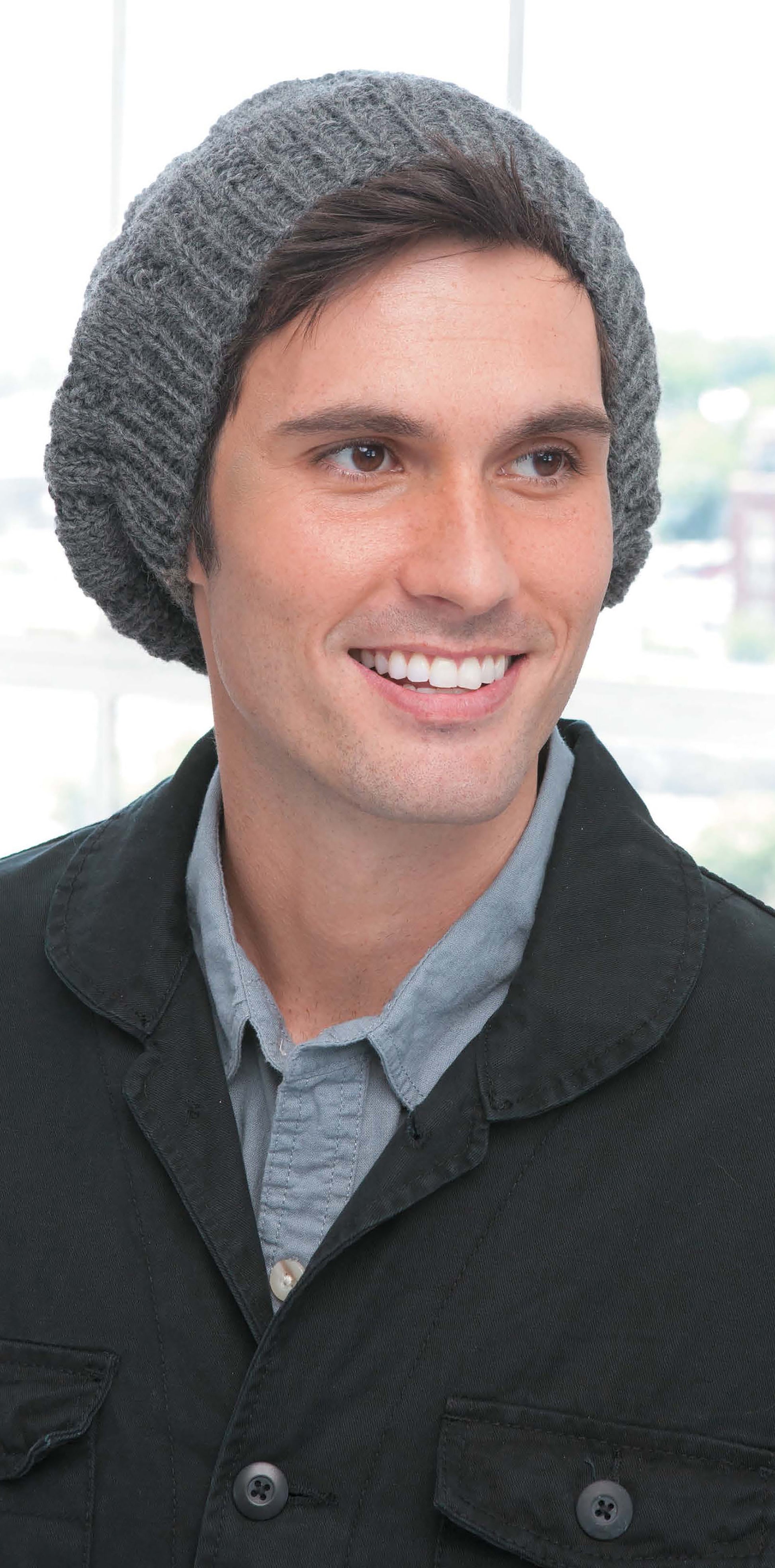 A smiling young man wears a Slouchy Beanie from Leisure Arts with a light gray button-up and black jacket, standing indoors against a bright, blurred background—showcasing winter fashion style.