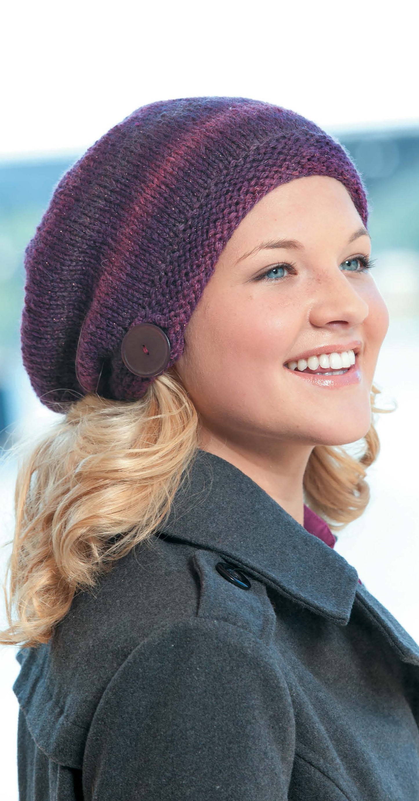 A smiling person with wavy blonde hair wears a Leisure Arts Slouchy Beanie in purple with a decorative button and a dark gray coat, highlighting cozy winter style from the Slouchy Beanies and Headwraps collection.