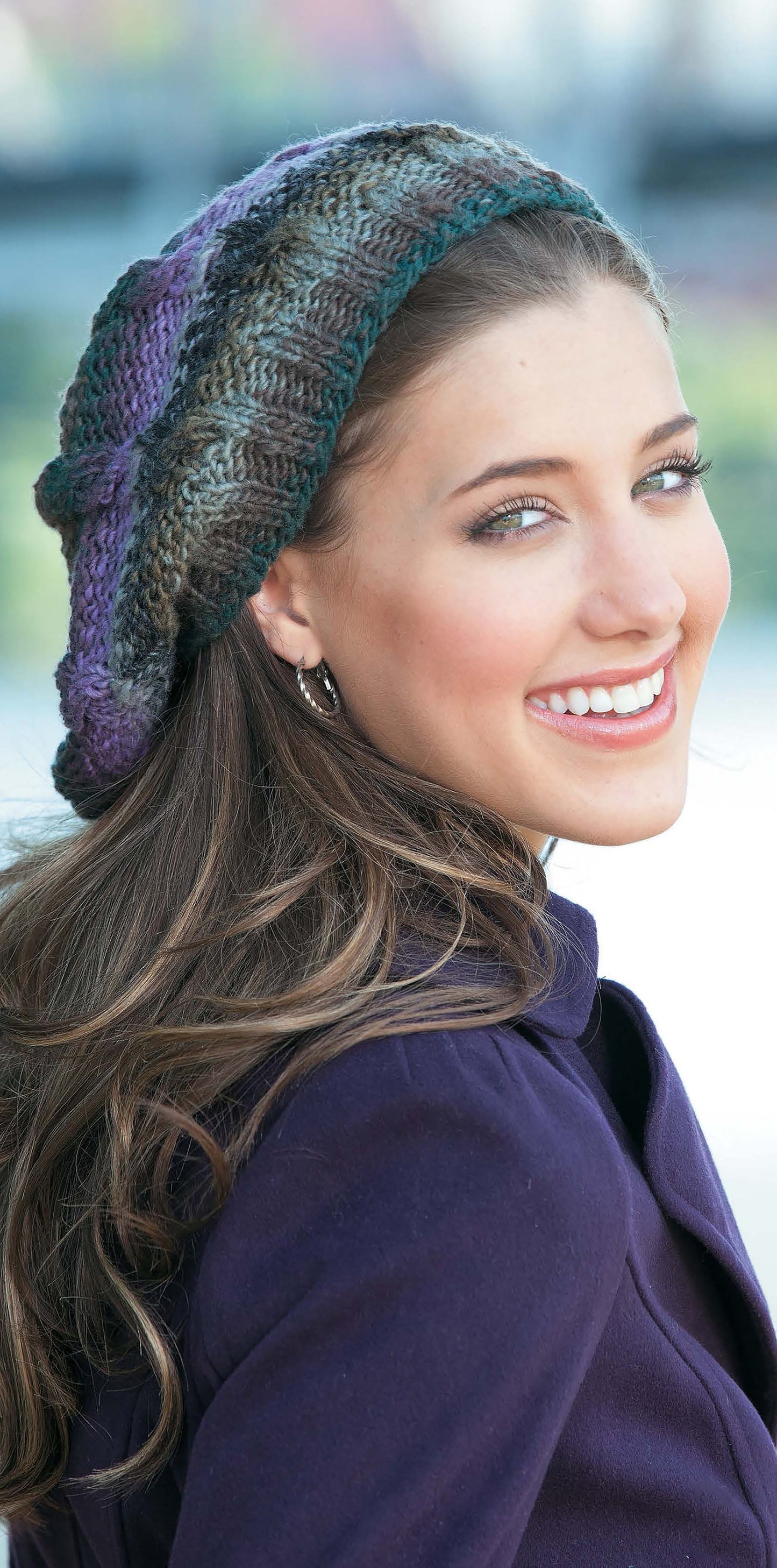 A young woman with long brown hair smiles over her shoulder in a multicolored Leisure Arts Slouchy Beanie, paired with a purple coat against a softly blurred background that evokes cozy winter style.