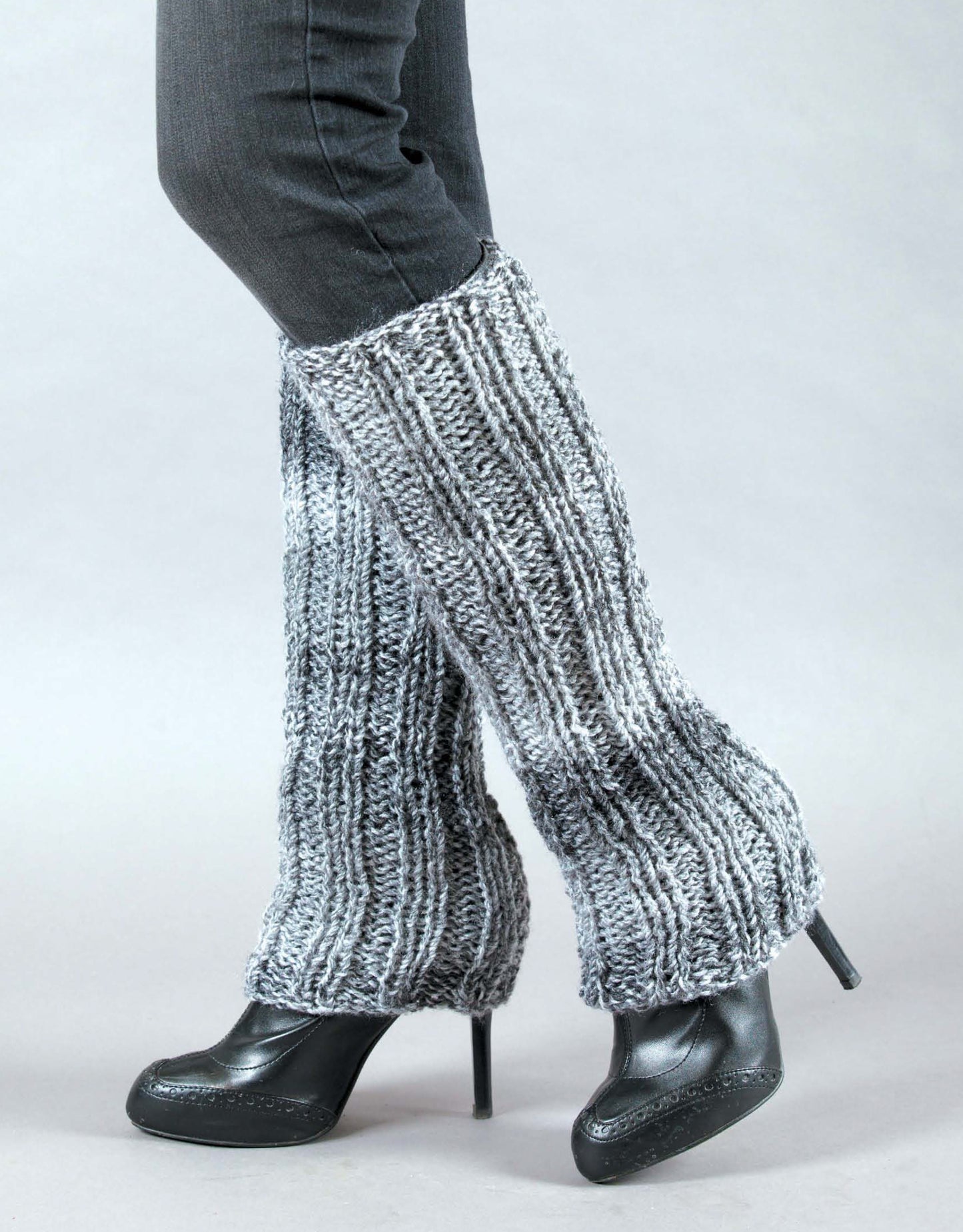 A person wears black heels, dark pants, and flared gray chunky-knit leg warmers—perfect inspiration for beginner knitters. Shown: "Learn to Knit, Now! Digital Download" by Leisure Arts. The background is plain and light-colored.