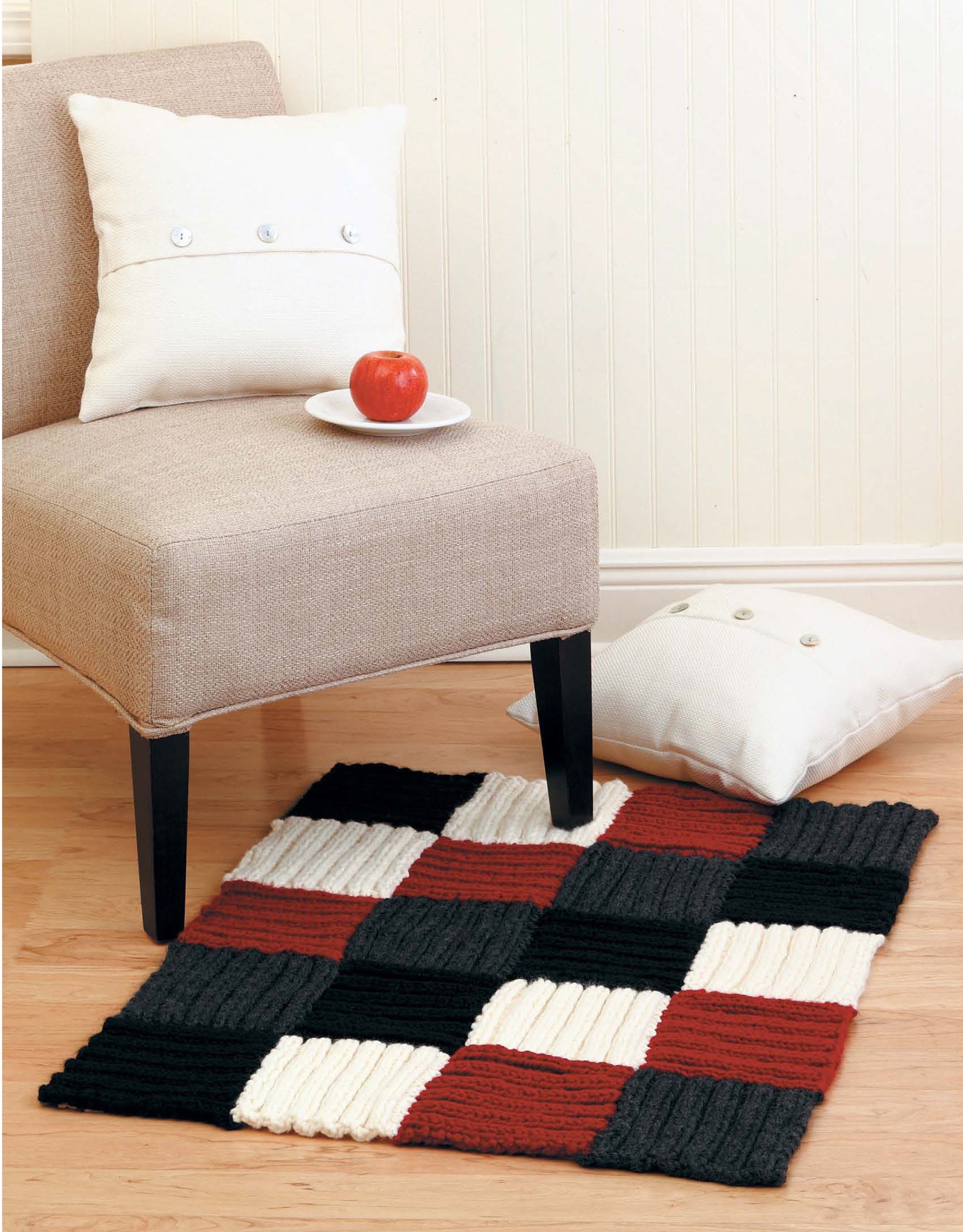 A beige chair with a white pillow and plate holding a red apple sits on light wood flooring. A second pillow is beside a knitted rug in black, red, and cream—great inspiration for beginner knitters using Leisure Arts' Learn to Knit, Now! Digital Download.