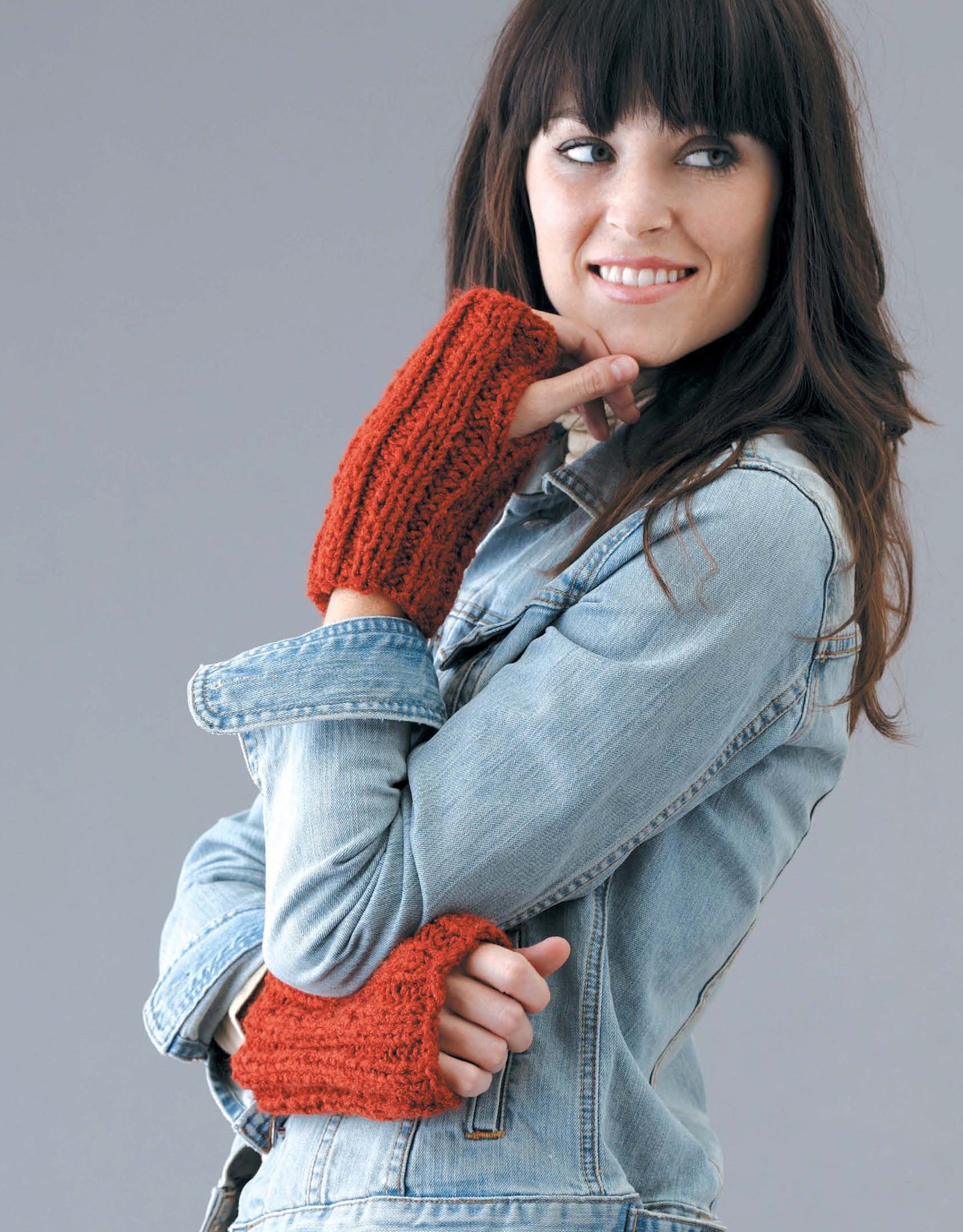 A woman with long dark hair smiles and looks to the side, wearing a light denim jacket and orange fingerless gloves made using Leisure Arts' Learn to Knit, Now! Digital Download—perfect for beginners. She stands against a plain gray background.