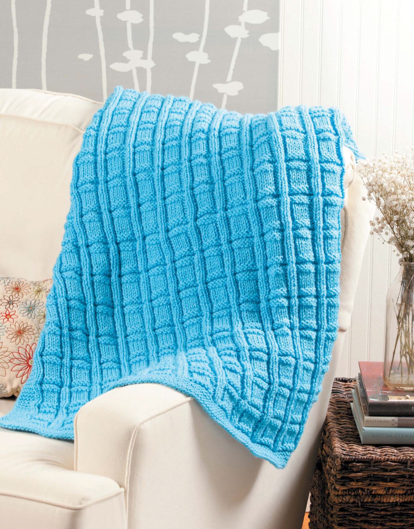 A bright blue textured blanket—great for beginner projects inspired by Leisure Arts' "Learn to Knit, Now!" digital download—is draped over a cream sofa with a floral pillow, next to a basket and vase of dried flowers.