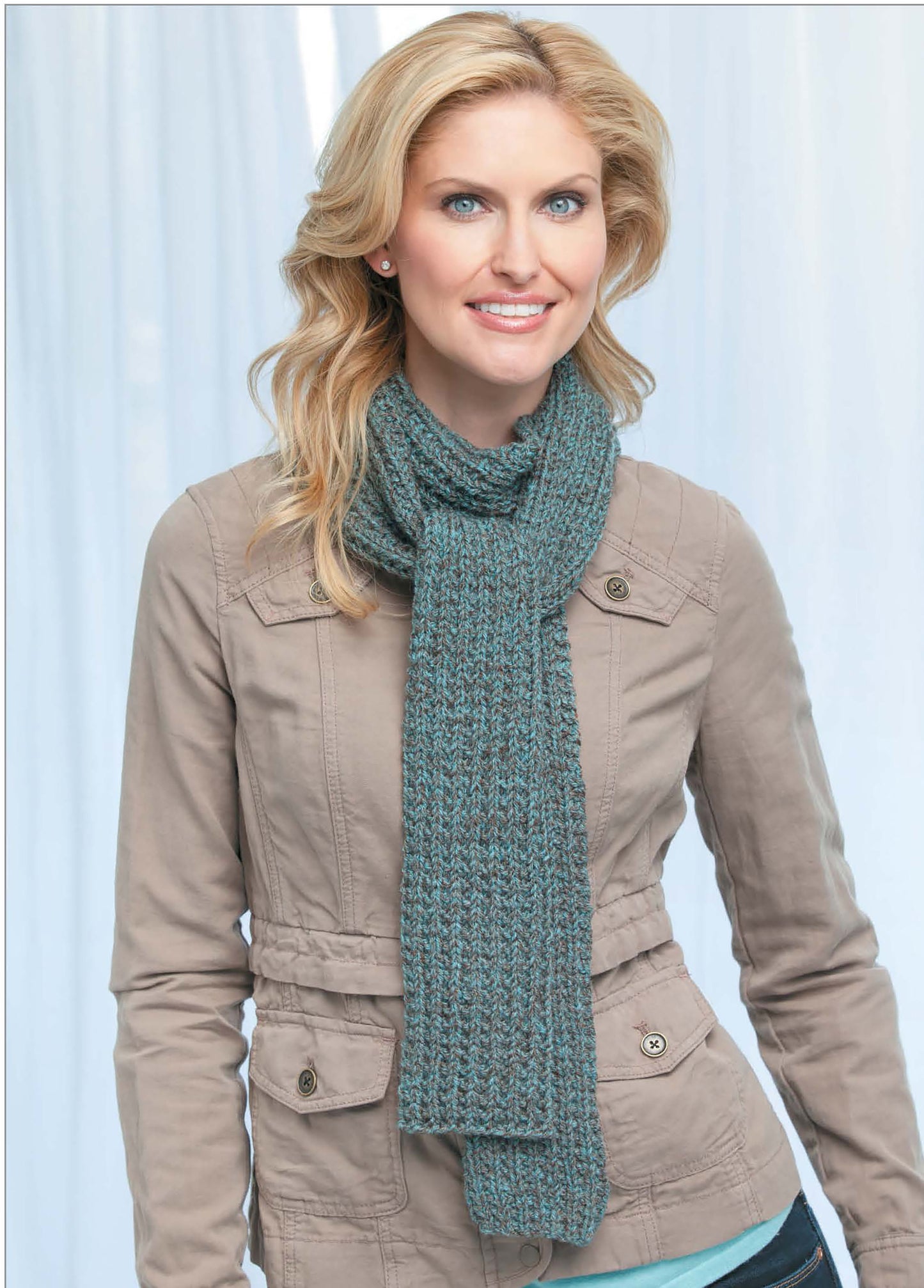 A smiling woman with wavy blonde hair wears a beige jacket and a blue scarf made using "Skinny Scarves to Knit" by Leisure Arts, standing in front of a softly blurred light background.