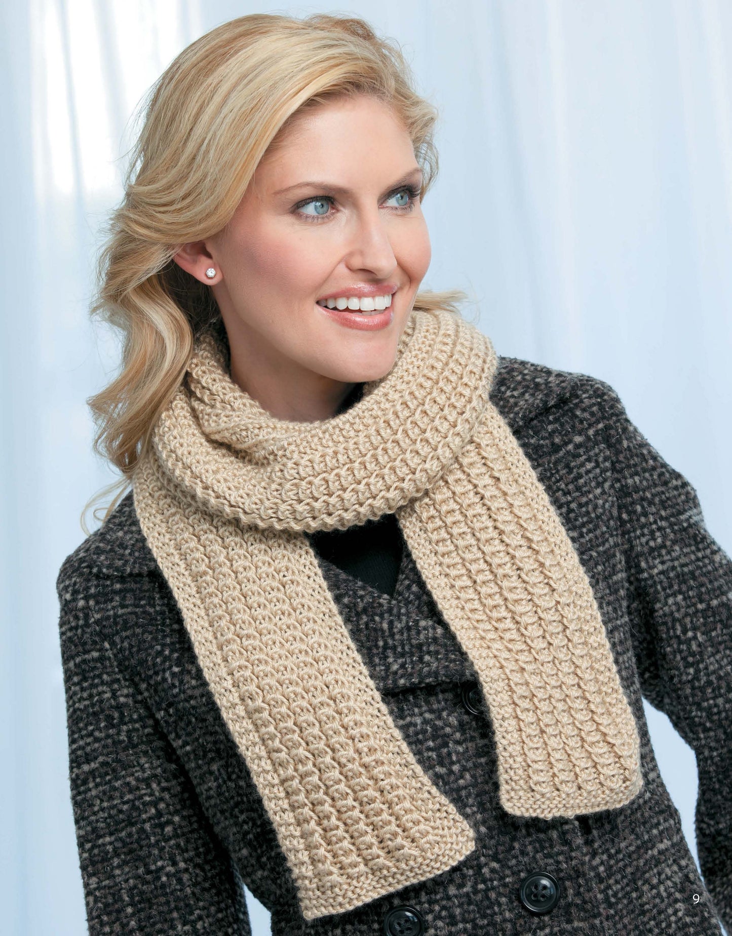 A smiling blonde woman wears a beige scarf from "Skinny Scarves to Knit" by Leisure Arts and a dark textured coat, standing against a light blue background.