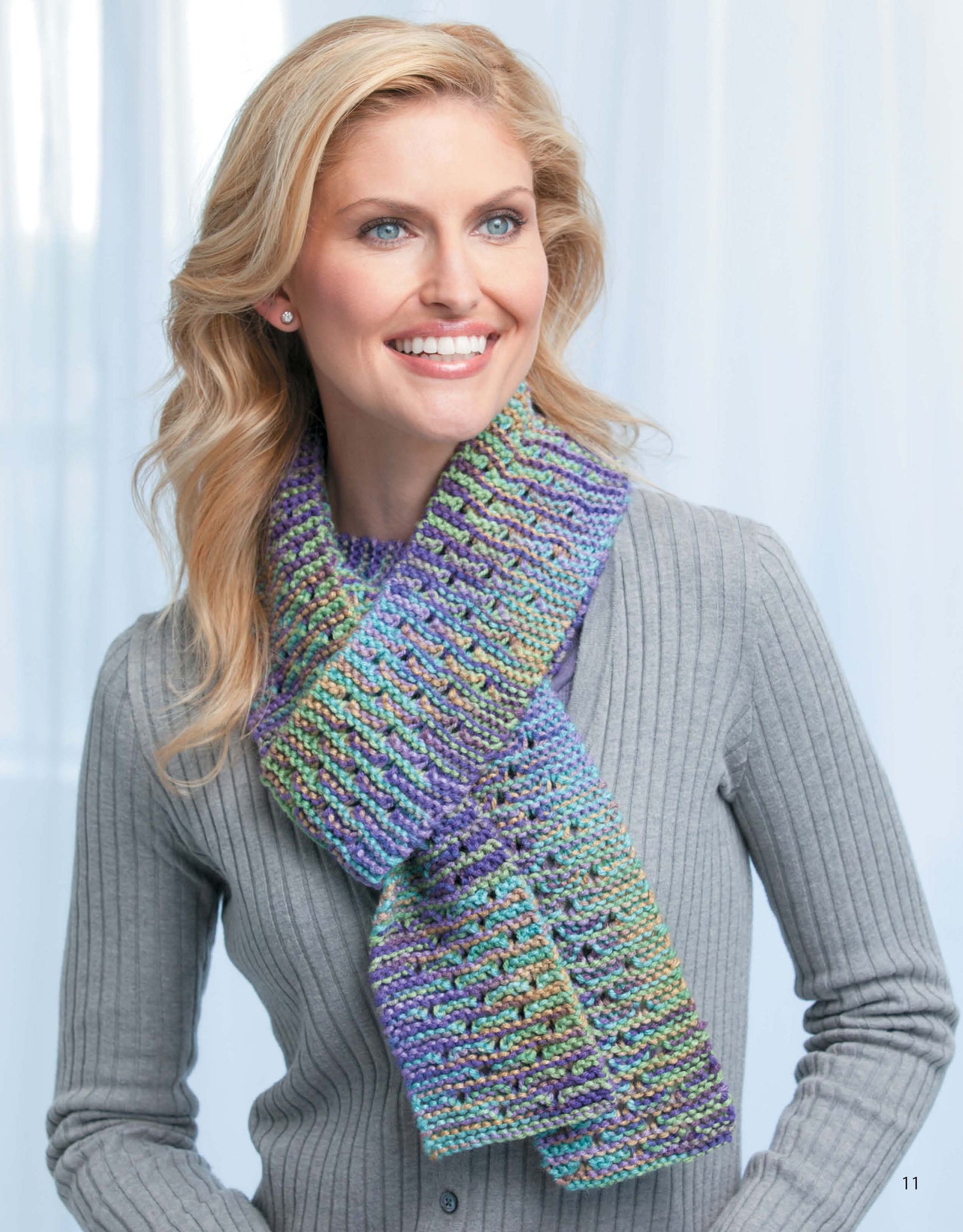 A smiling woman with wavy blonde hair models a gray ribbed sweater and a vibrant knit scarf from Leisure Arts' "Skinny Scarves to Knit," ideal for anyone looking for easy knitting patterns, against a softly blurred light backdrop.