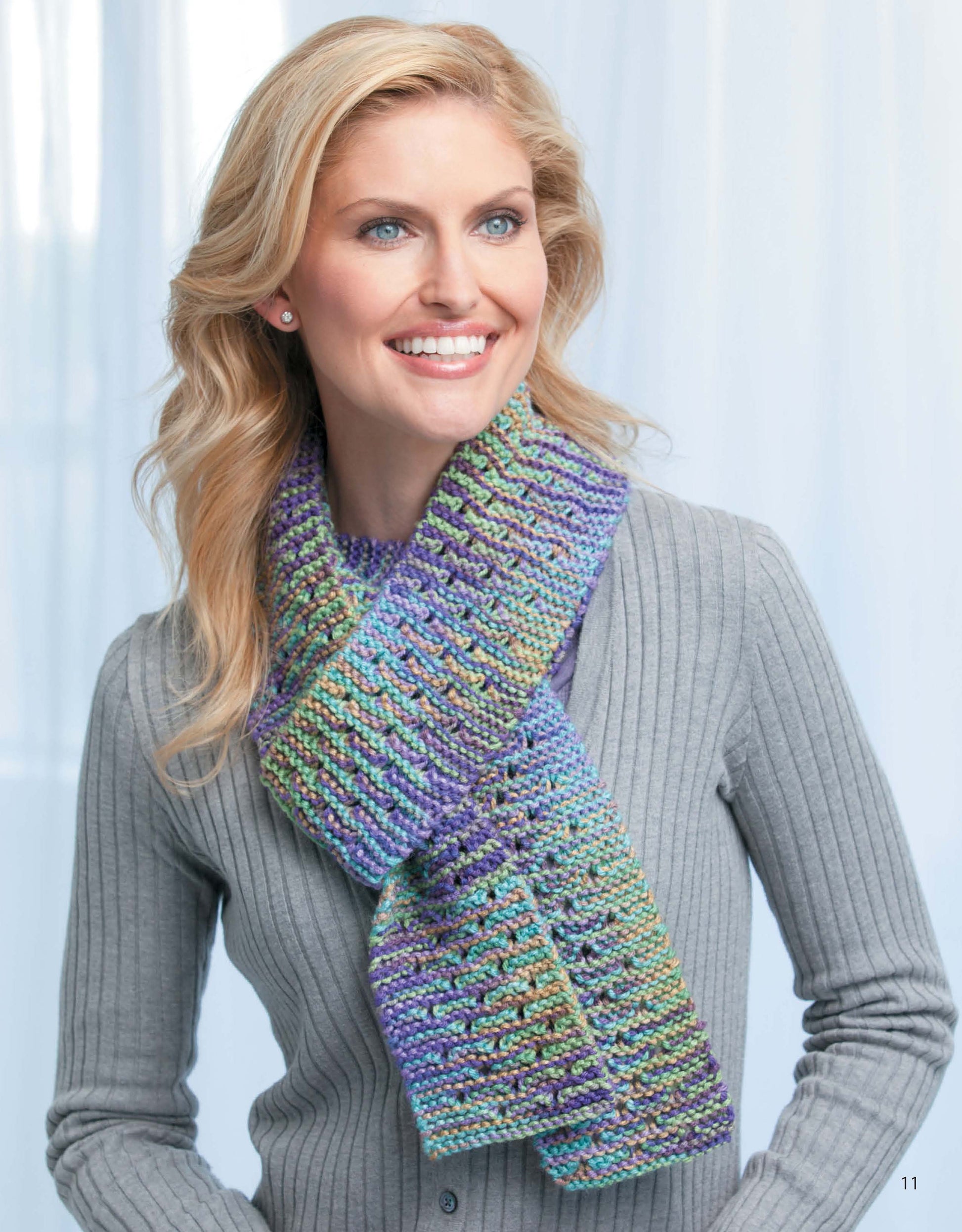 A smiling woman with wavy blonde hair models a gray ribbed sweater and a vibrant knit scarf from Leisure Arts' "Skinny Scarves to Knit," ideal for anyone looking for easy knitting patterns, against a softly blurred light backdrop.