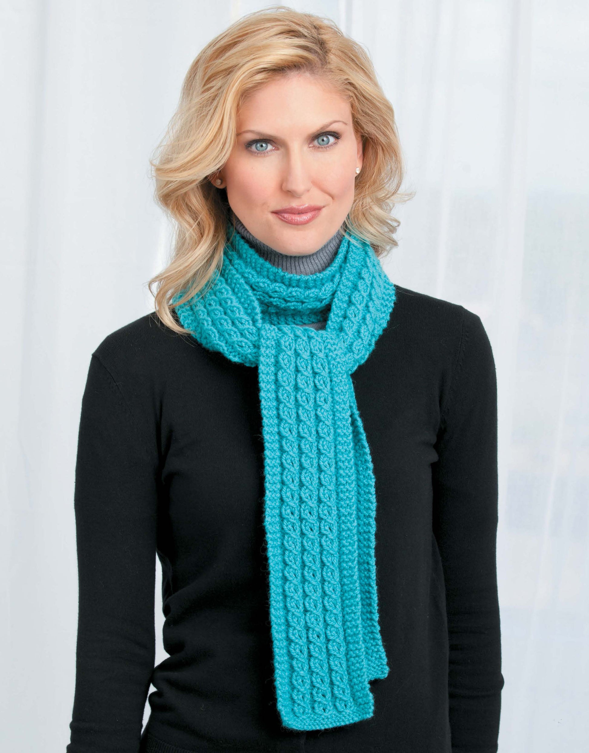 A woman with blonde hair wears a black sweater and a bright turquoise scarf made from Leisure Arts’ "Skinny Scarves to Knit," standing before a white curtain and looking at the camera with a neutral expression.