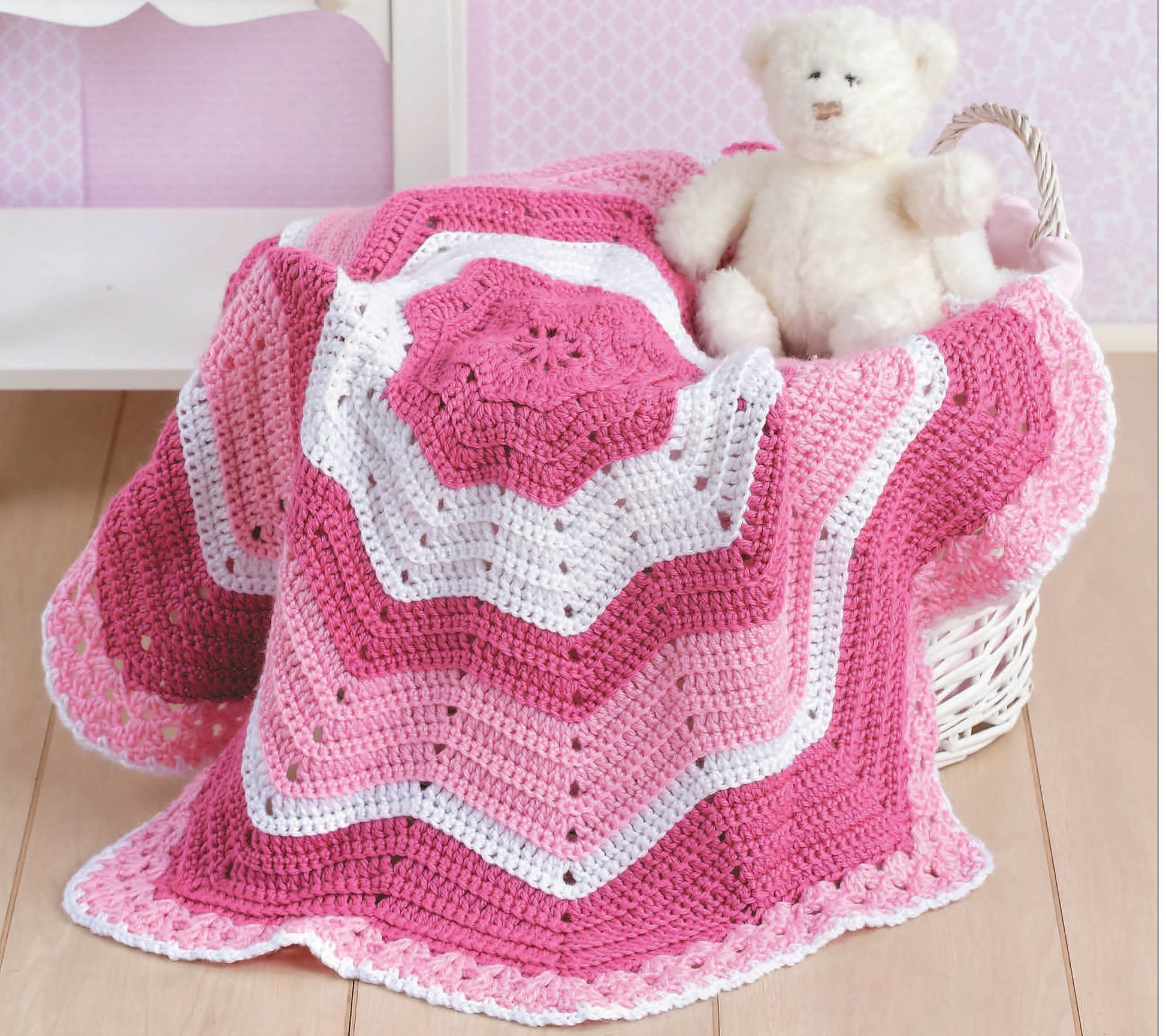 A white wicker basket holds a stuffed white teddy bear and a crocheted pink, white, and magenta star-shaped baby blanket made with patterns from "Baby Afghans" by Leisure Arts, displayed on a light wood floor with pink wallpaper in the background.