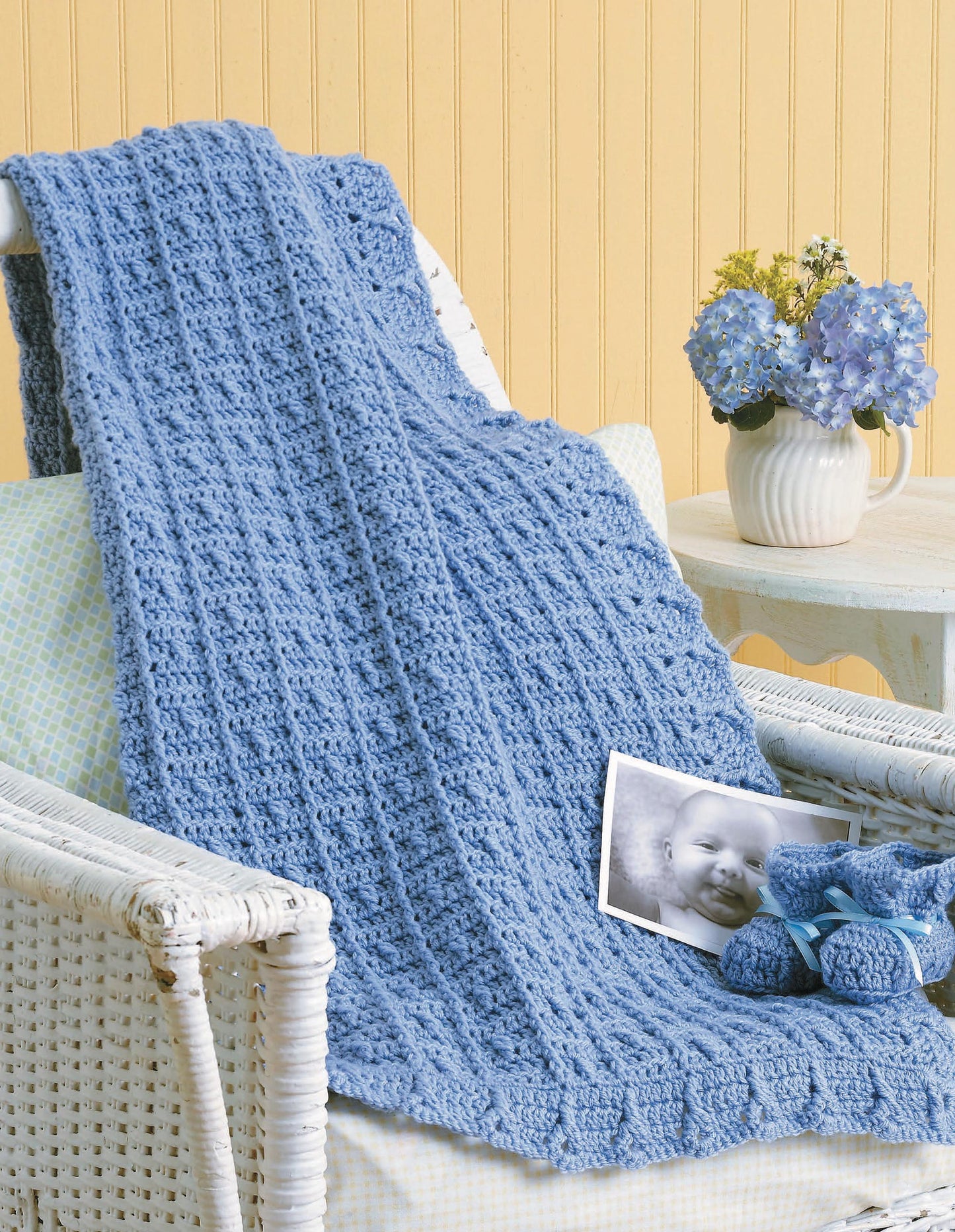 A cozy blue crochet baby blanket from Leisure Arts’ "Baby Afghans — 8 Cozy Wrap Patterns for Your Special Wee One" is draped over a wicker chair, paired with blue booties and cheerful decor—ideal for crochet pattern enthusiasts.