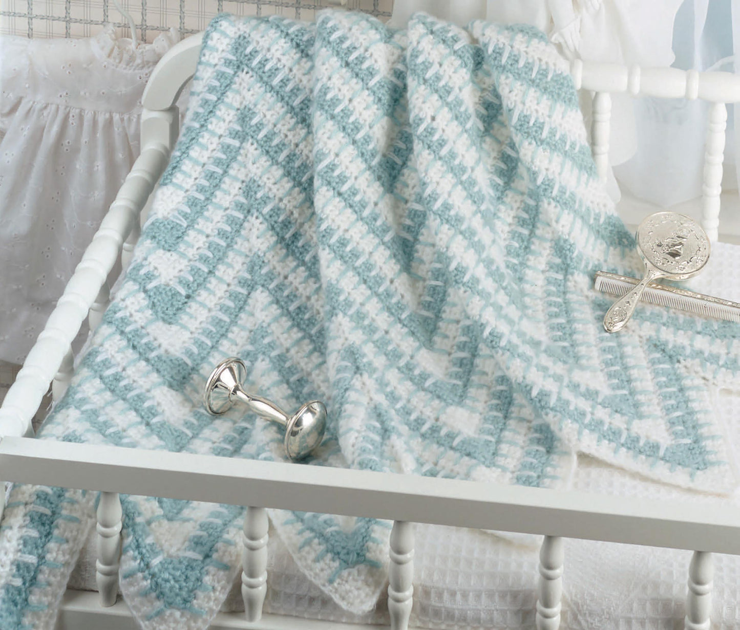 A Leisure Arts "Baby Afghans" soft blue and white chevron knit blanket, inspired by beginner crochet patterns, is draped over a white crib with silver baby rattles on the bedding—perfect for your special wee one.
