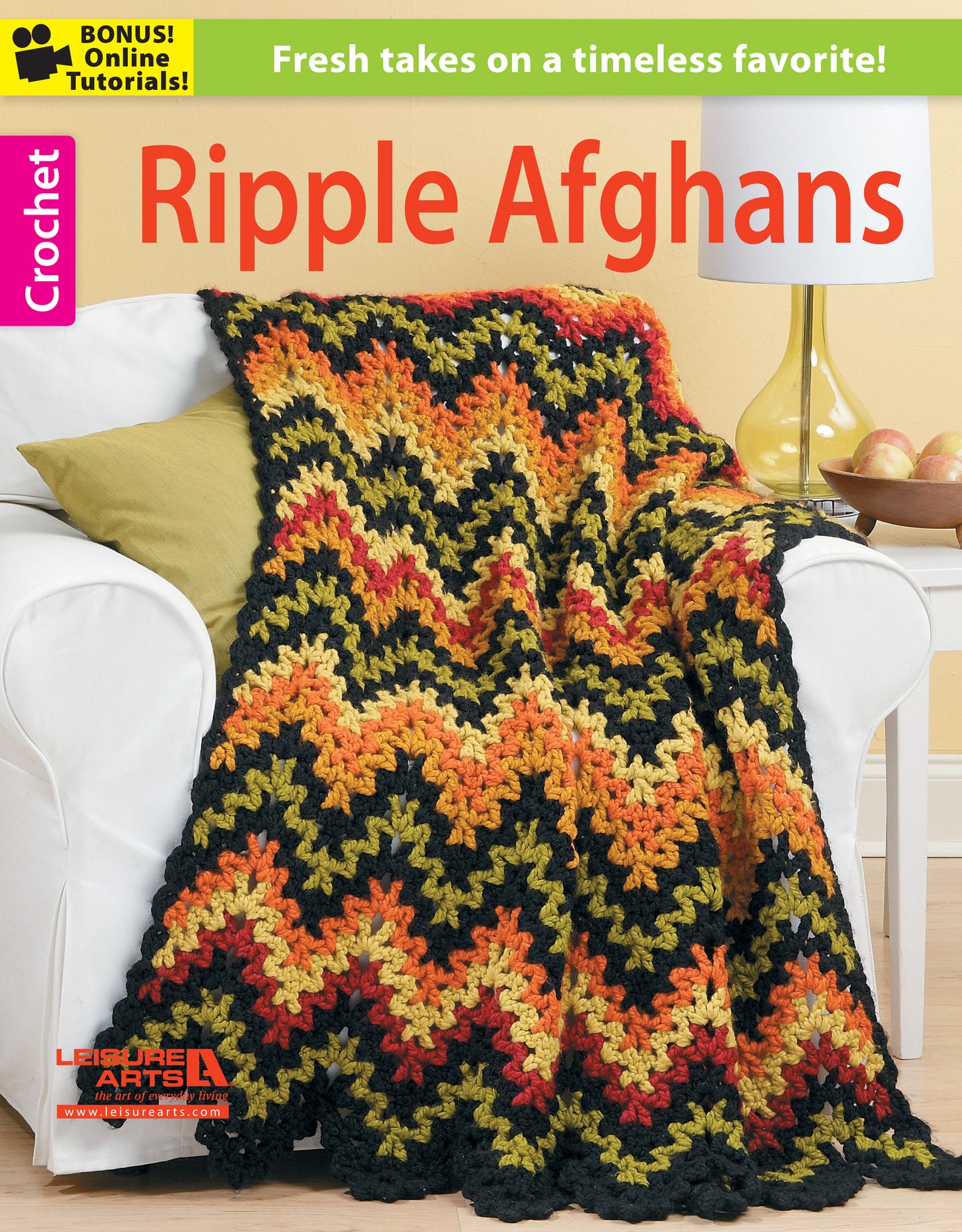 A vibrant Crochet Ripple Afghan from Leisure Arts in yellow, orange, red, and black is draped over a white chair near a yellow lamp and apples. Text: Crochet Ripple Afghans by Leisure Arts—fresh takes on a timeless favorite!.