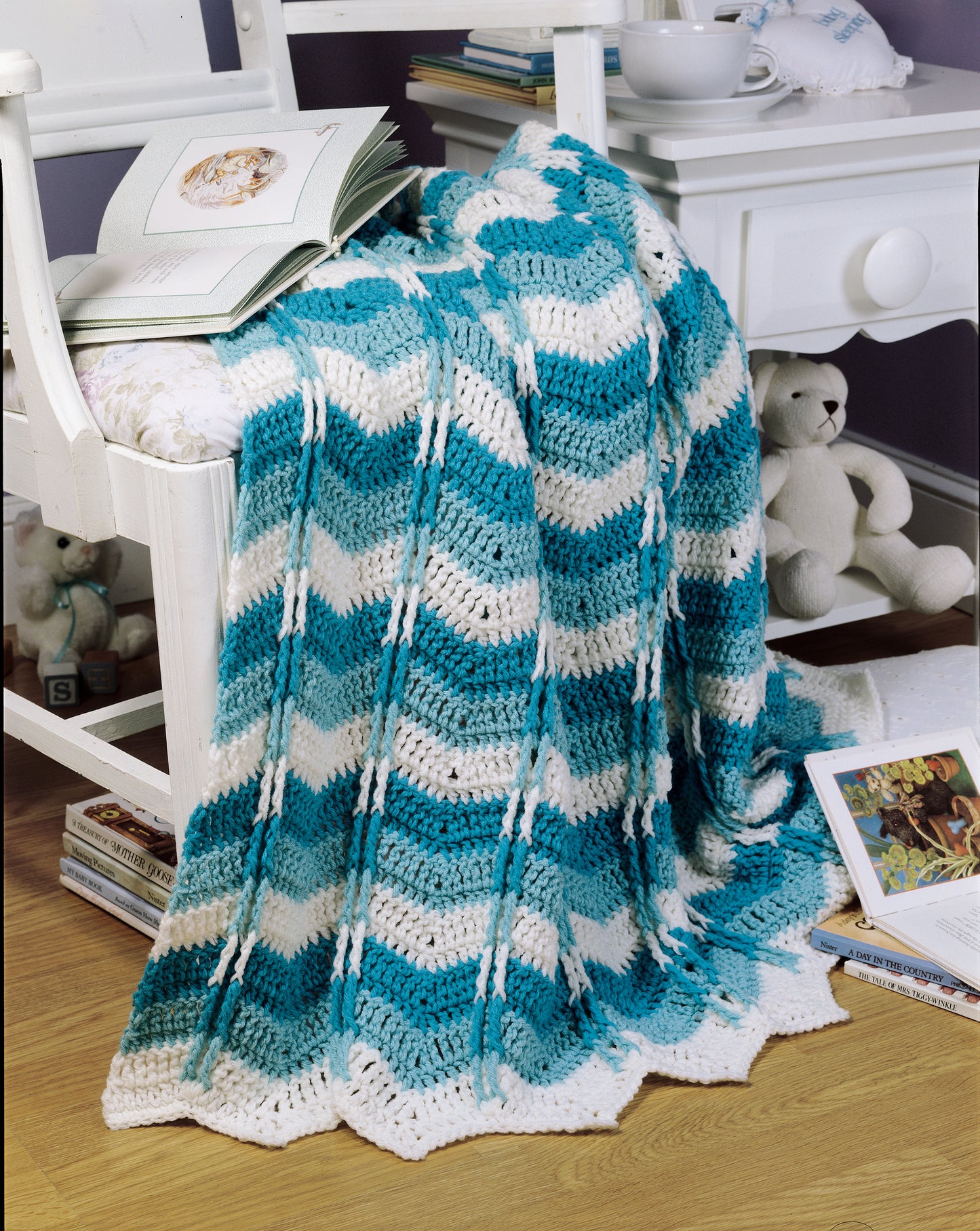 A blue and white chevron-patterned "Crochet Ripple Afghan" by Leisure Arts is draped over a cushioned white chair next to a nightstand, with children’s books and a white teddy bear on the wooden floor nearby.