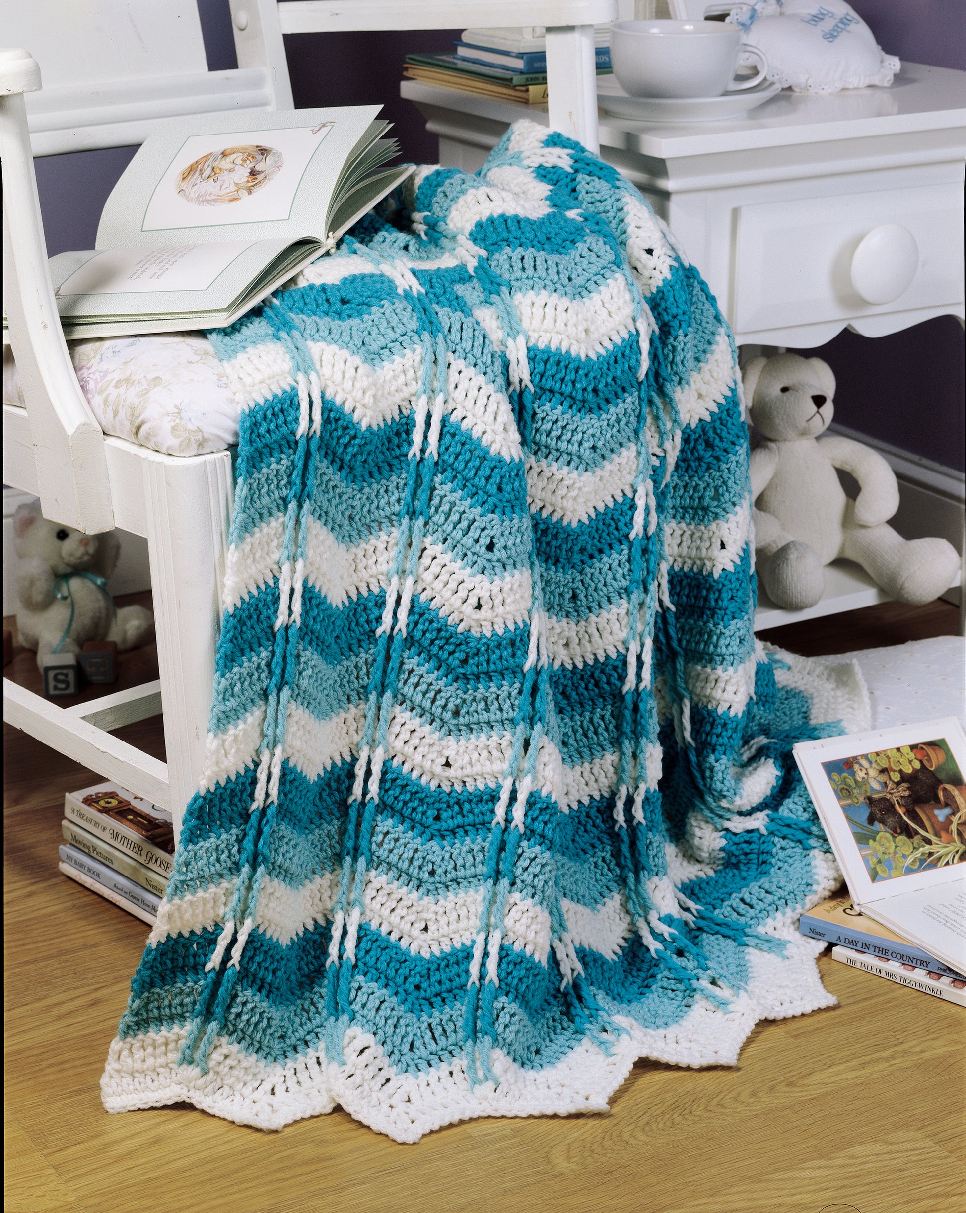 A blue and white chevron-patterned "Crochet Ripple Afghan" by Leisure Arts is draped over a cushioned white chair next to a nightstand, with children’s books and a white teddy bear on the wooden floor nearby.