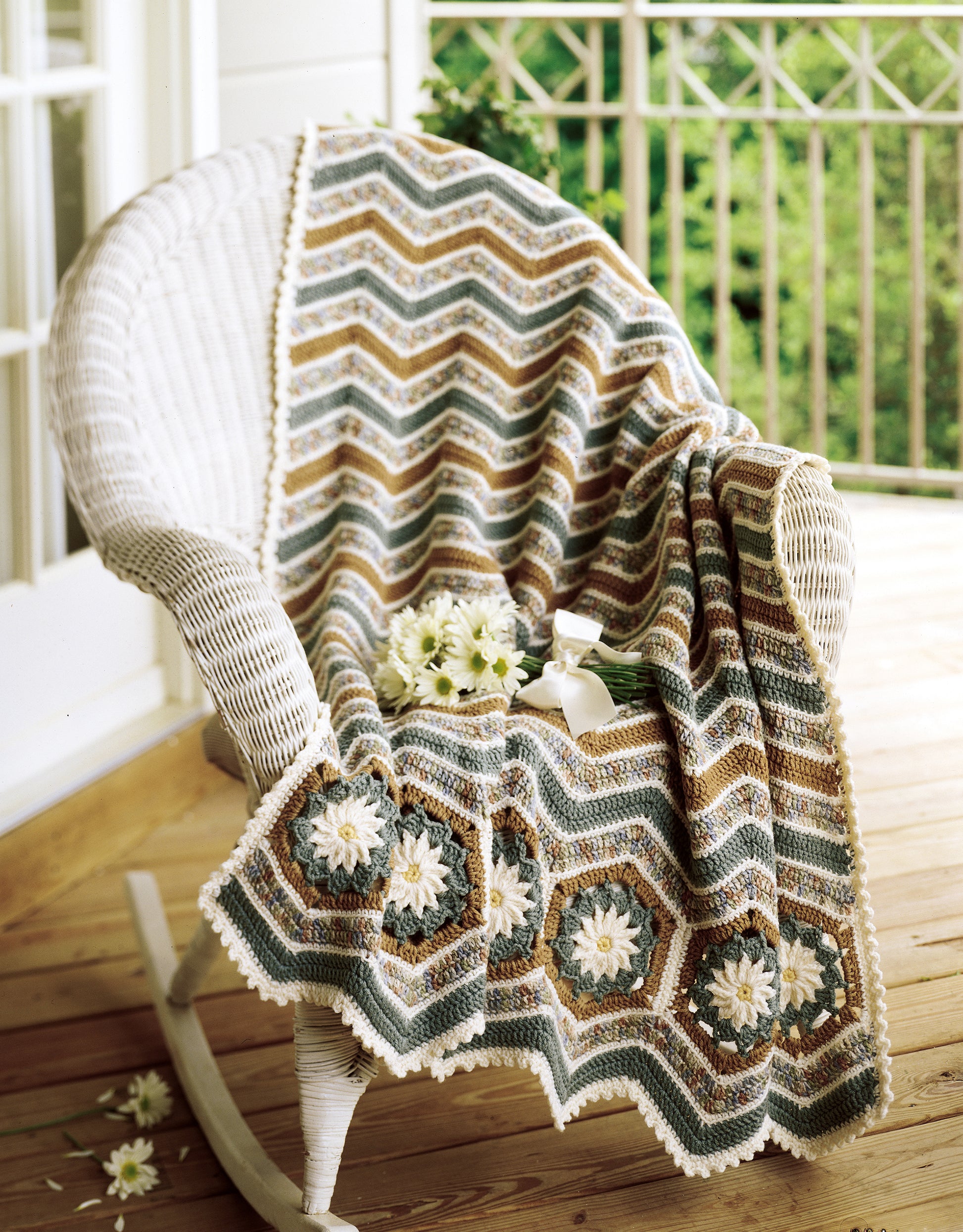 A white wicker rocking chair on a wooden porch displays the "Crochet Ripple Afghans" by Leisure Arts, made in green, brown, and cream worsted weight yarn with daisy motifs; a bouquet of white daisies rests on the blanket.