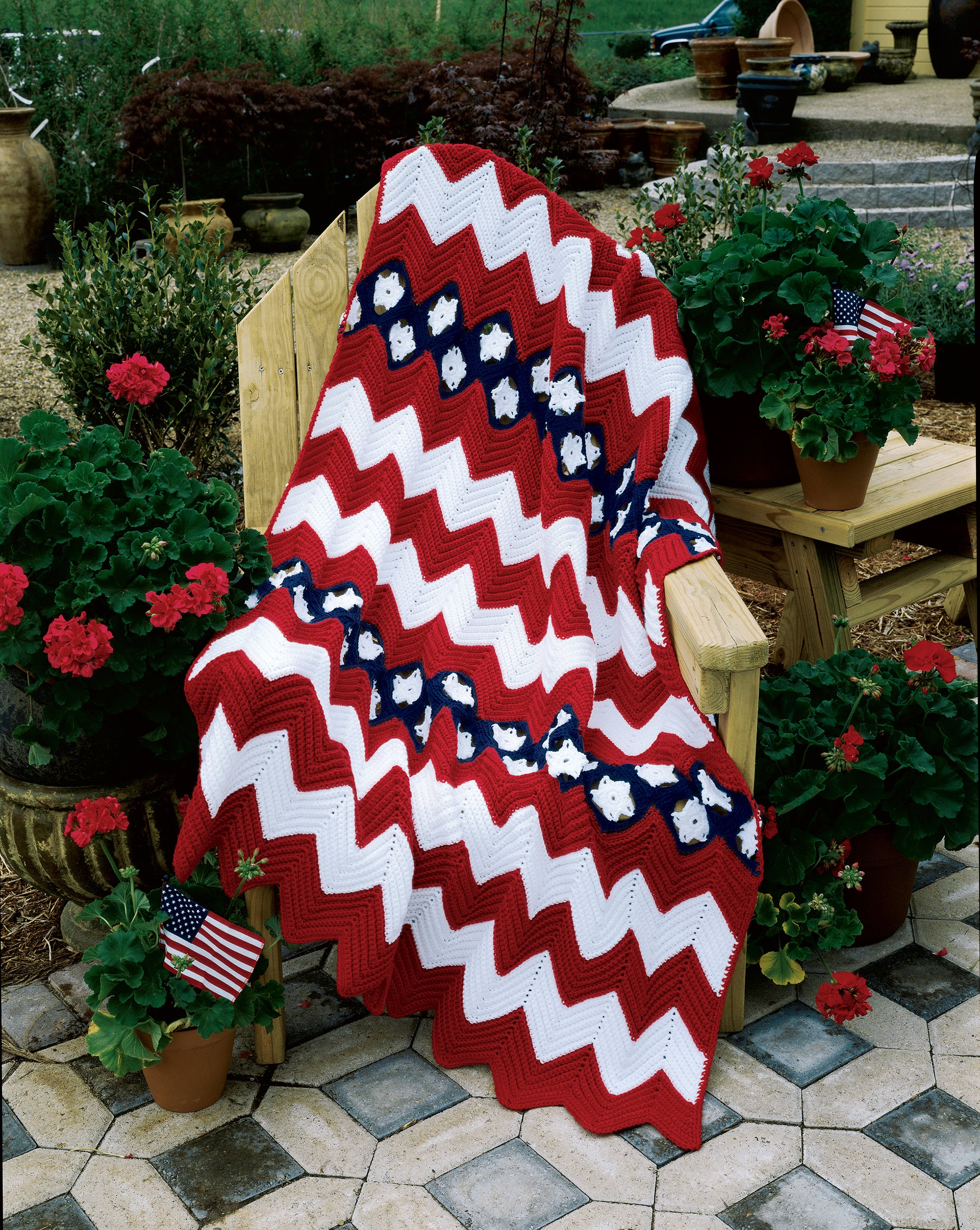The Crochet Ripple Afghans by Leisure Arts, featuring a red, white, and blue zigzag design with star motifs, is draped over a wooden chair surrounded by potted geraniums and American flags on a stone patio.