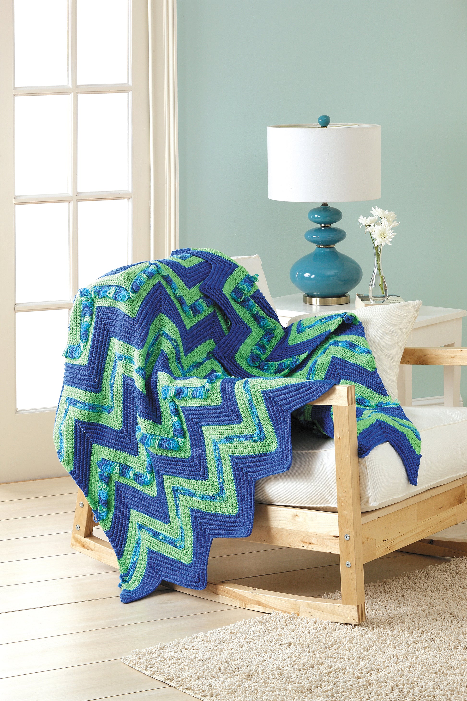 A cozy chair by the window features a Leisure Arts Crochet Ripple Afghans blanket draped over it, paired with white cushions, a blue lamp, and a small vase of white flowers on the nearby side table.
