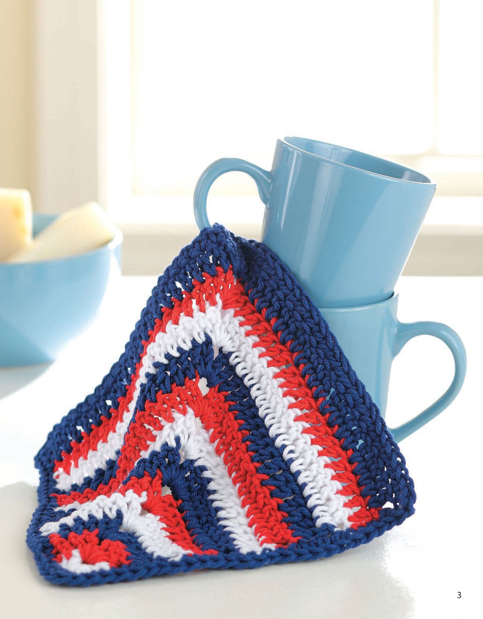 Two blue mugs are stacked on a counter, topped with a red, white, and blue crocheted dishcloth from Leisure Arts' "Dishcloths—A Super Collection for Home or Gifts! Digital Download." A light blue bowl of sliced fruit brightens this cozy kitchen scene.