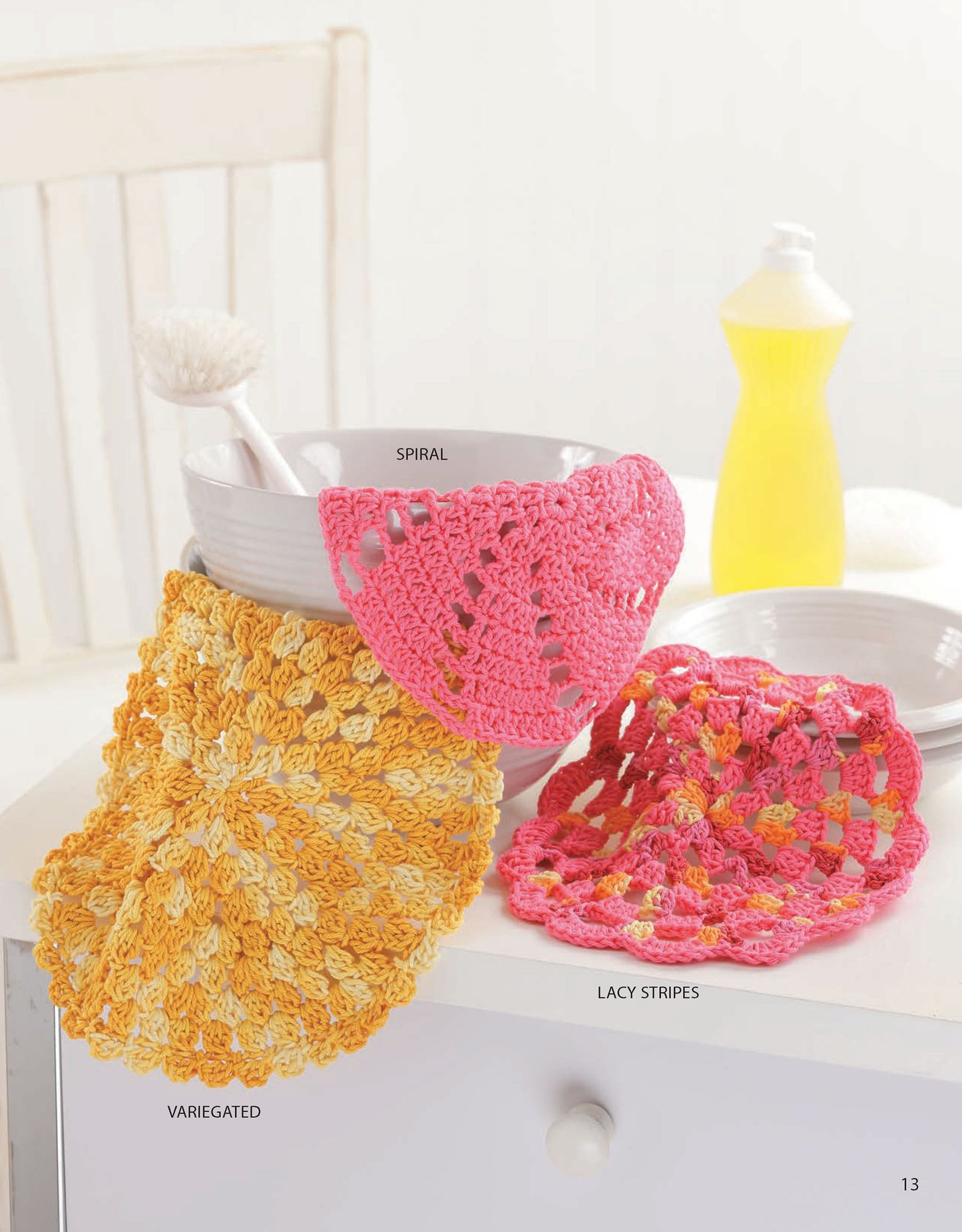 A white bowl with a dish brush sits on a table, displaying three colorful crocheted dishcloths from Leisure Arts' "Dishcloths—A Super Collection for Home or Gifts! Digital Download." A yellow dish soap bottle adds a cheerful touch in the background.