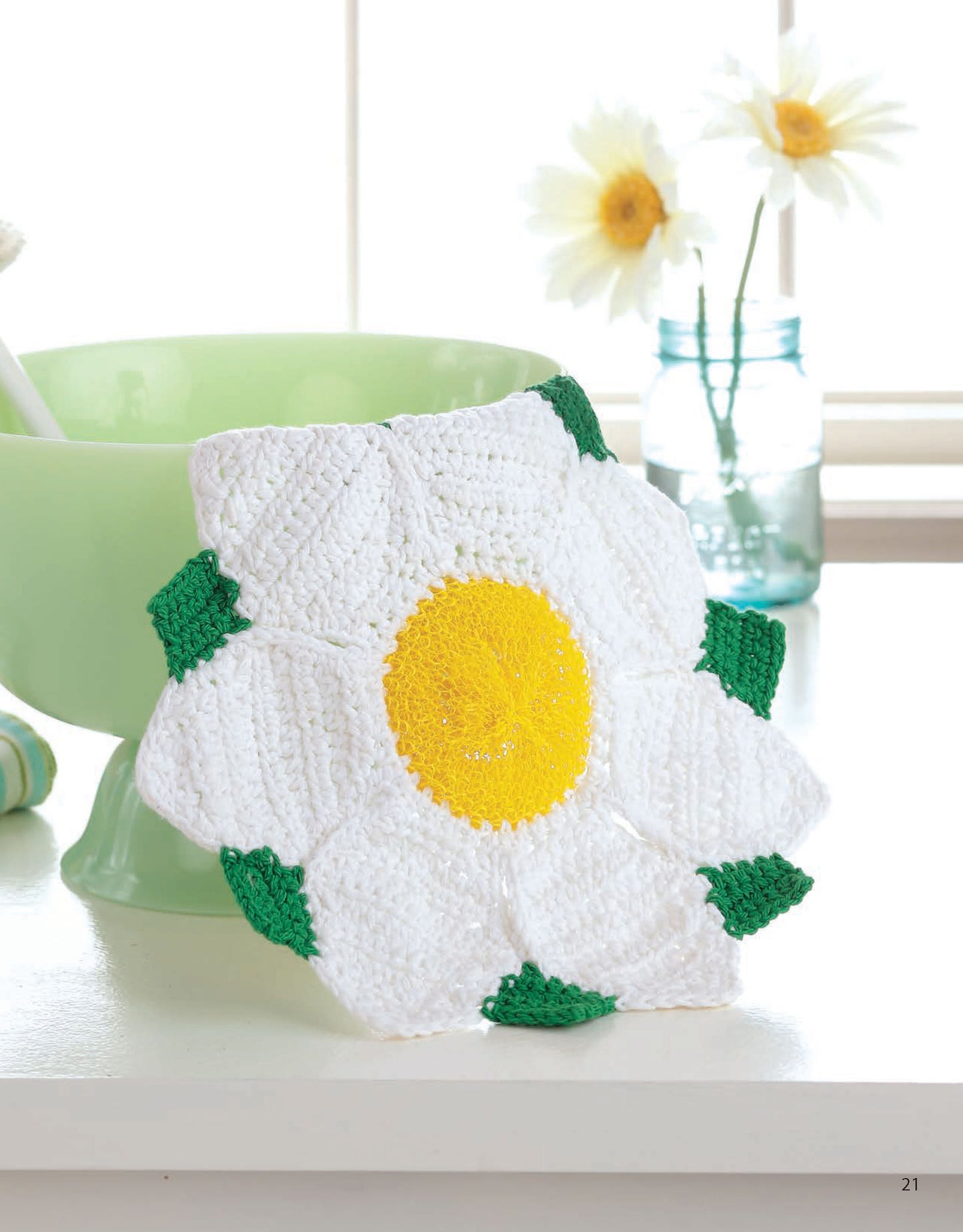 A white daisy with a yellow center and green leaves from Leisure Arts’ "Dishcloths—A Super Collection for Home or Gifts! Digital Download" rests by a light green bowl, while two daisies in a blue glass jar brighten the windowsill.