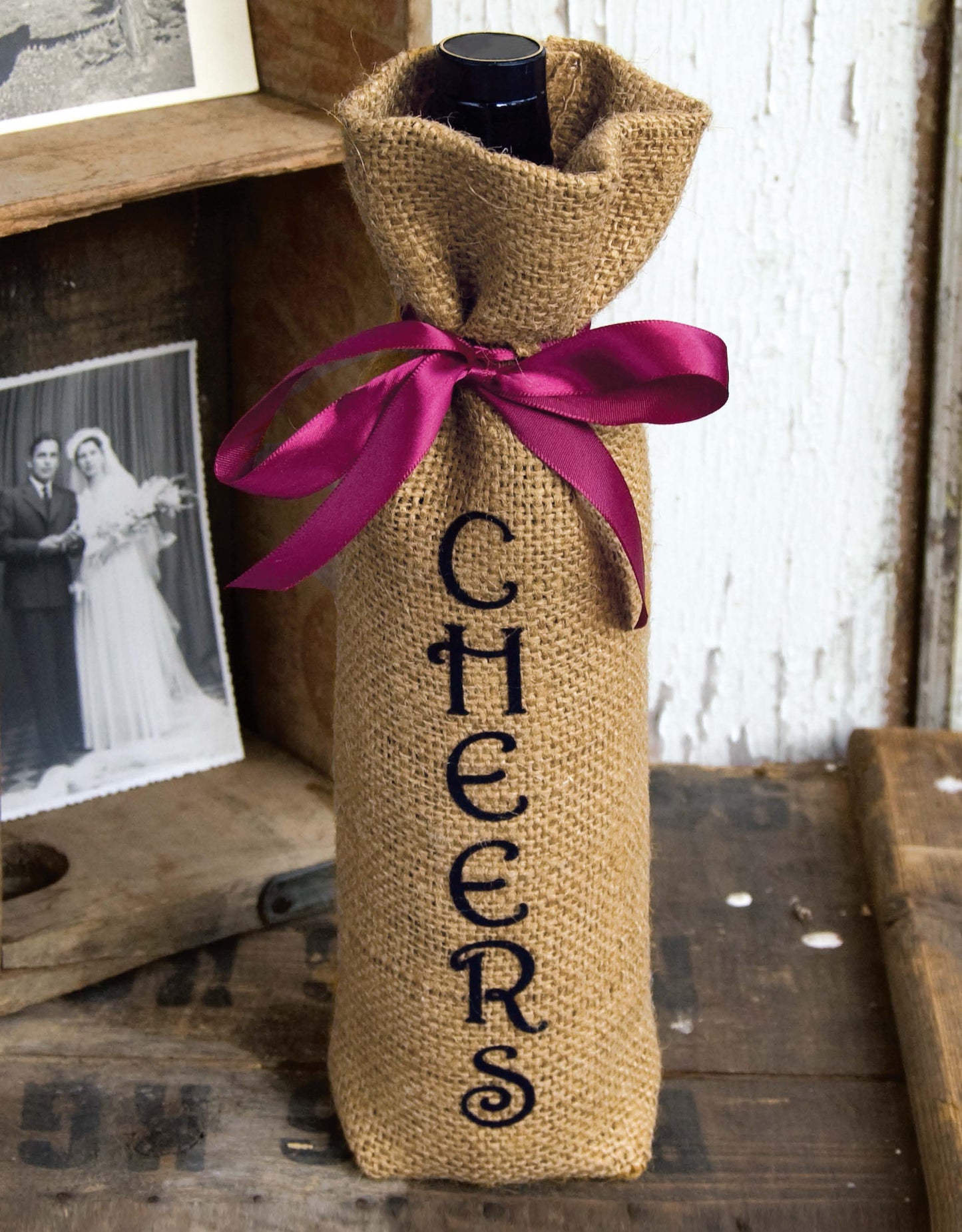 A wine bottle wrapped in burlap with “CHEERS” printed vertically and tied with a dark purple ribbon shows creative decorating from Leisure Arts’ Decorate with Burlap, displayed on a rustic wood surface near black-and-white photos.