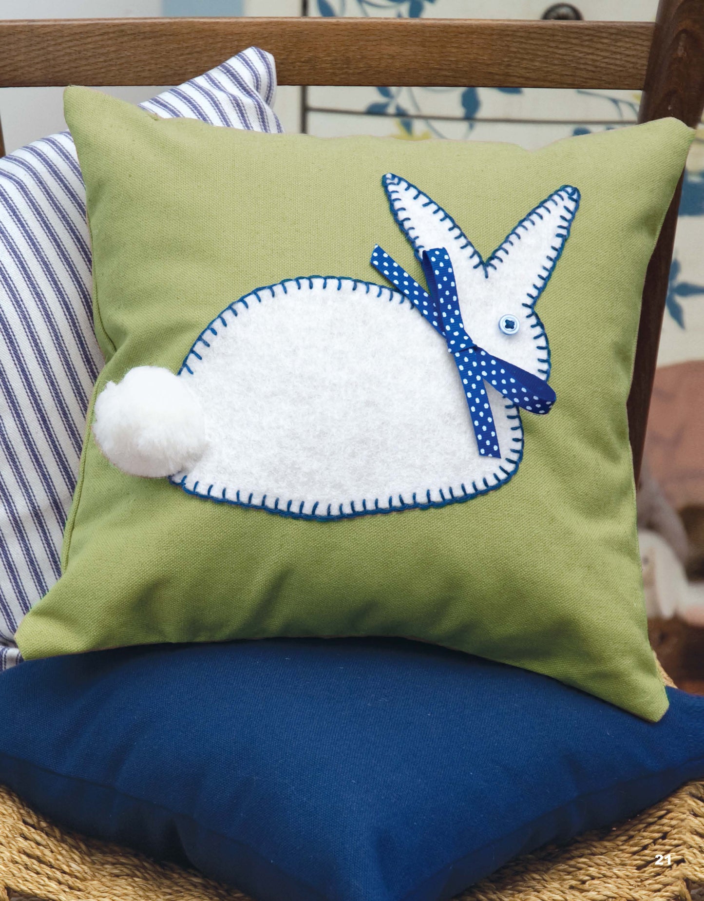 The "Decorate with Burlap" kit by Leisure Arts features a green cushion with a handmade felt white rabbit appliqué, blue blanket stitch edging, polka-dot bow, button eye, and pom-pom tail to add charm to burlap décor on a wooden chair.