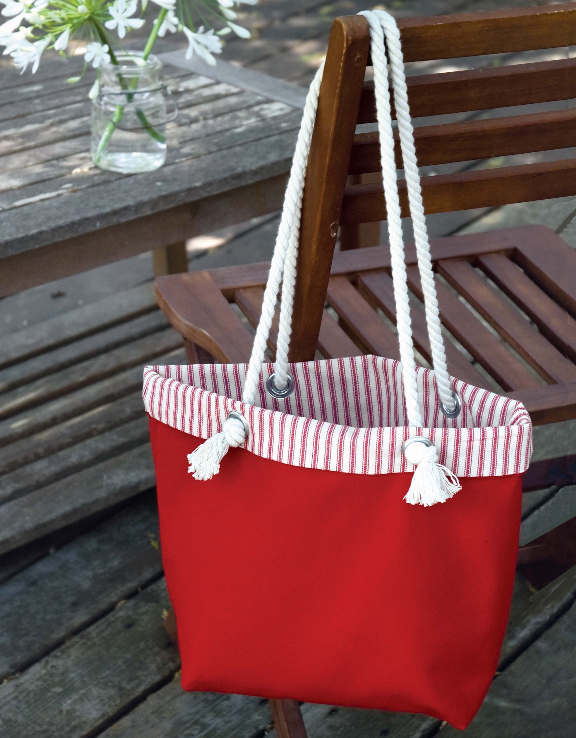 A red tote bag with white rope handles and striped lining sits on a wooden chair, next to the book "Decorate with Burlap" by Leisure Arts, perfect for inspiring simple no-sew burlap projects.