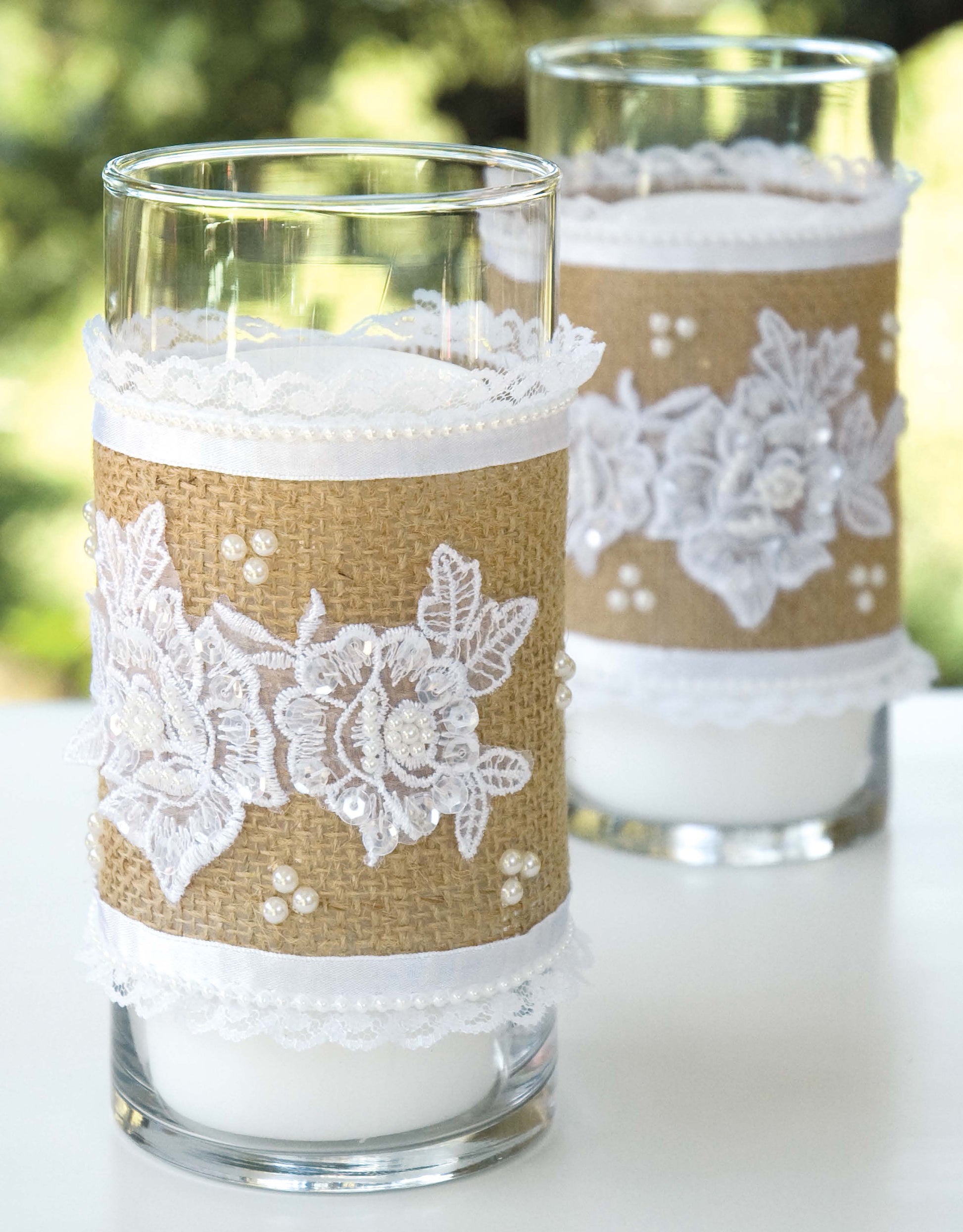 The "Decorate with Burlap" candle holders by Leisure Arts feature glass wrapped in burlap, white lace, and floral appliqués—ideal for rustic décor or no-sew burlap inspiration. Shown on a white surface with a blurred green background.