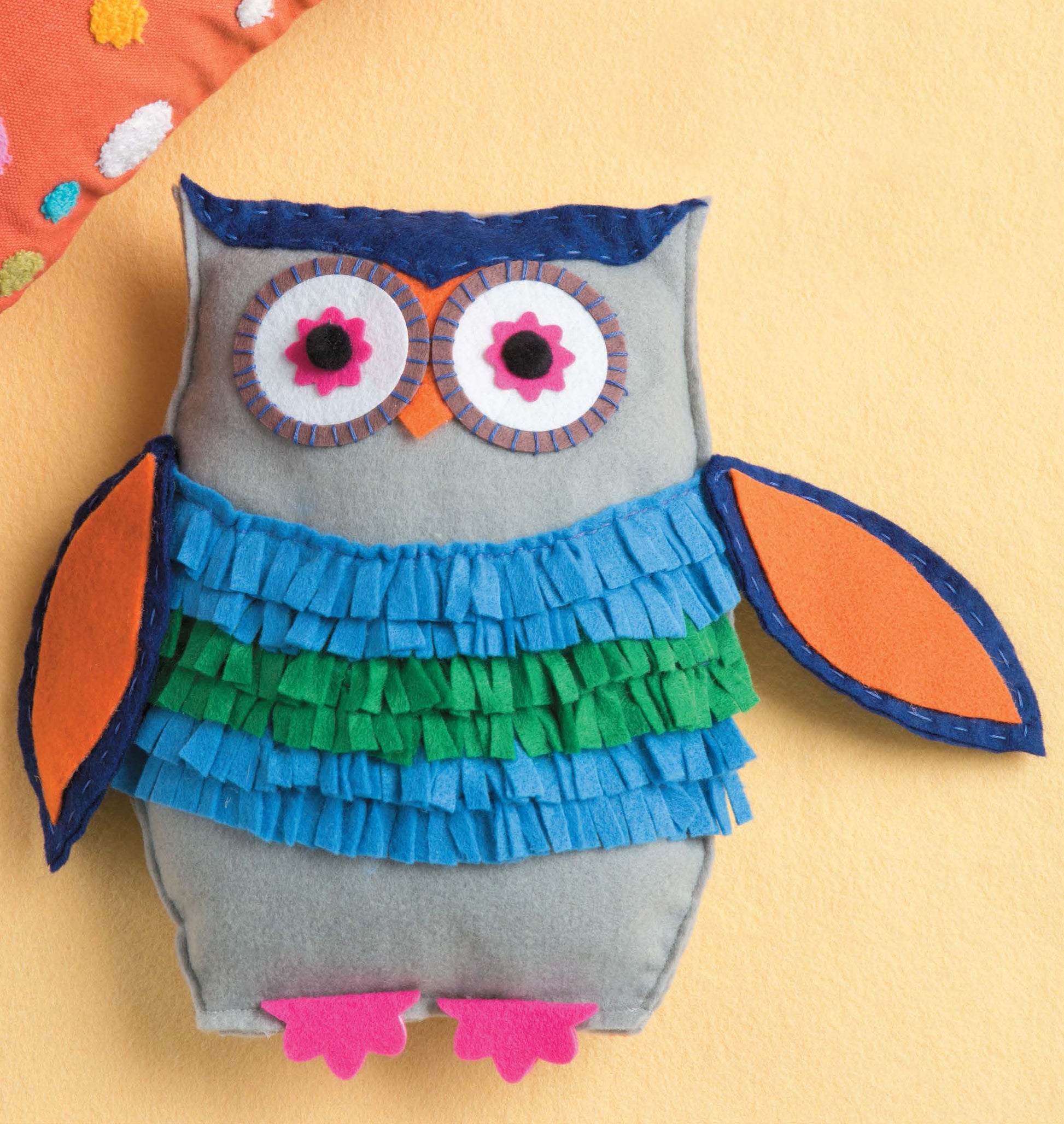 The Leisure Arts "Crafting with Felt" owl plush toy features a green and blue fringed belly, orange wings, pink feet, and large pink and black eyes—ideal as kids’ toys or unique home decor—shown beside a colorful dotted pillow.