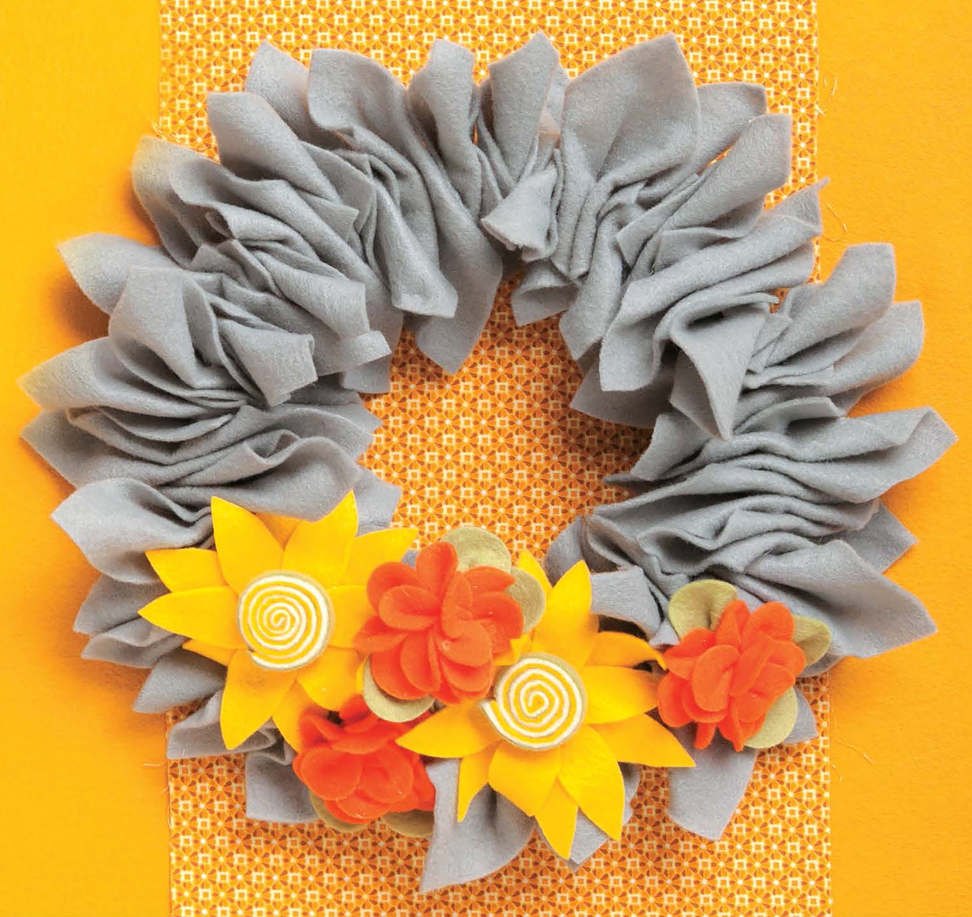 Crafting with Felt by Leisure Arts features a gray, ruffled fabric wreath adorned with yellow and orange felt flowers, displayed on an orange patterned background—an ideal piece for cheerful home decor.