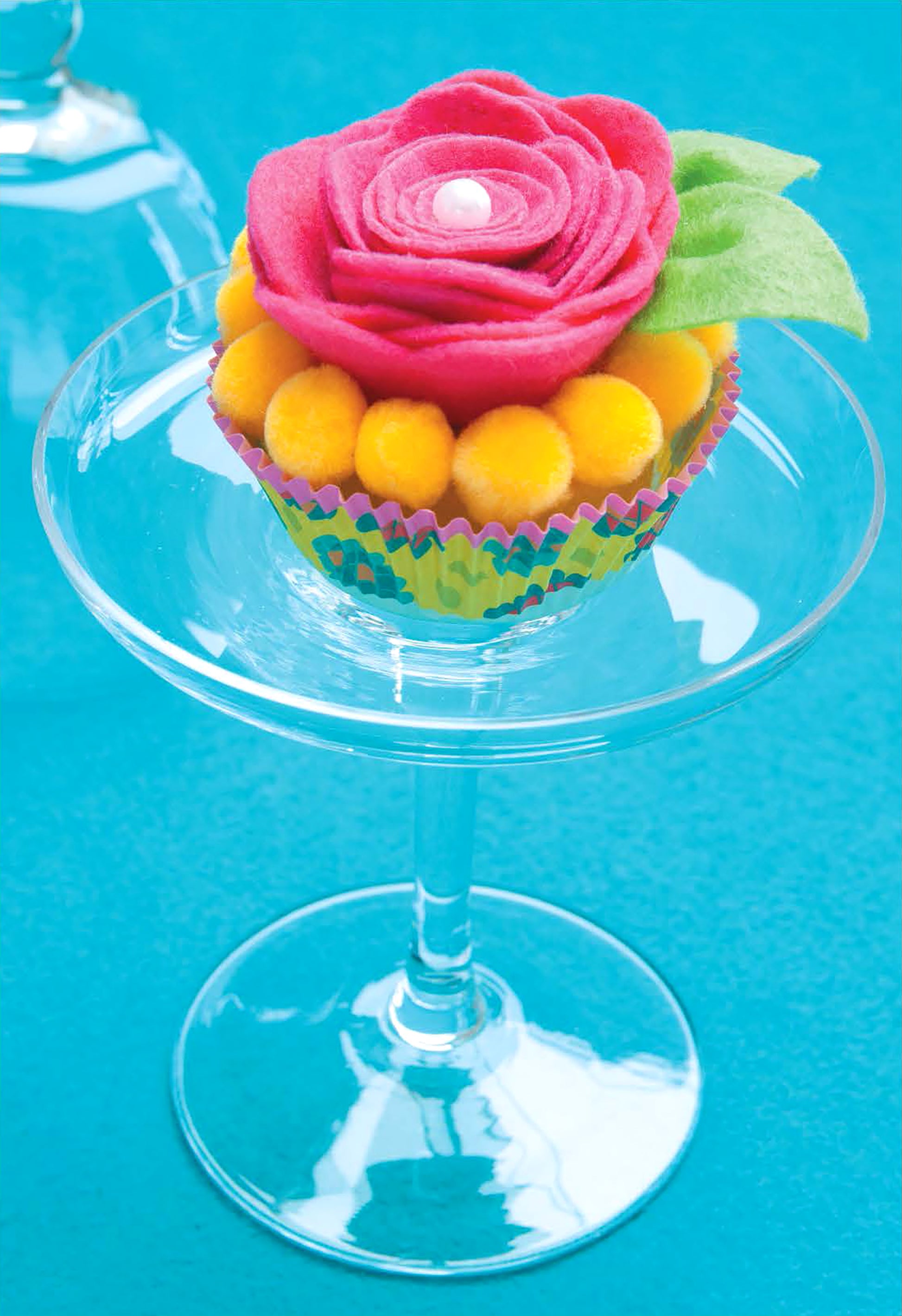 “Crafting with Felt” by Leisure Arts is displayed on a glass dessert stand, adorned with bright yellow accents and set against a vivid blue background—a whimsical home decor idea for creative spaces.