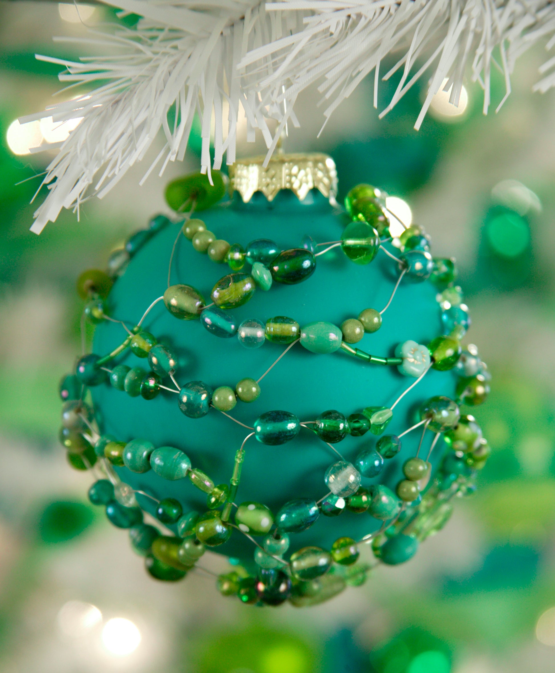 The Holiday Ornaments Digital Download by Leisure Arts features a teal ornament with spiraling green beads of various sizes, displayed on a white tree branch against a blurred green bokeh background.